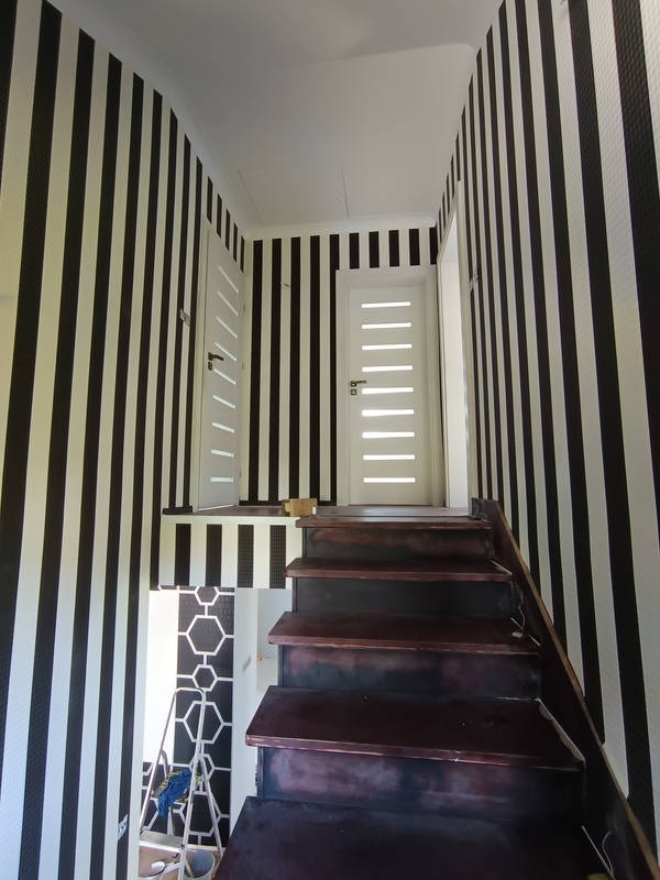 Klatka schodowa z tapetą w czarno-białe pasy; widoczne drewniane schody w ciemnym odcieniu oraz drabina w tle z elementami dekoracyjnymi na ścianie.