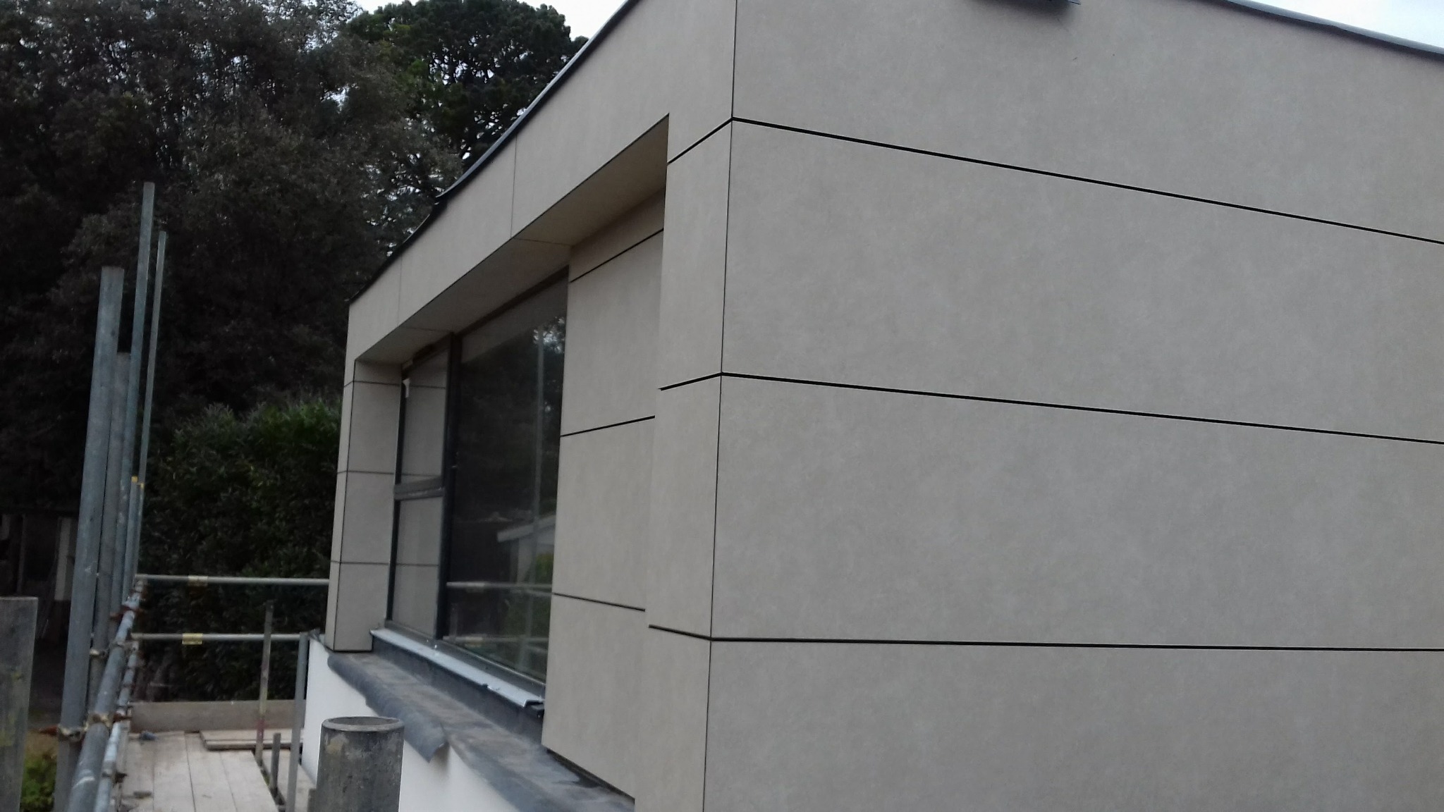 Fragment budynku z nowoczesną elewacją z paneli w odcieniach szarości, widoczne rusztowania sugerujące prace w toku.