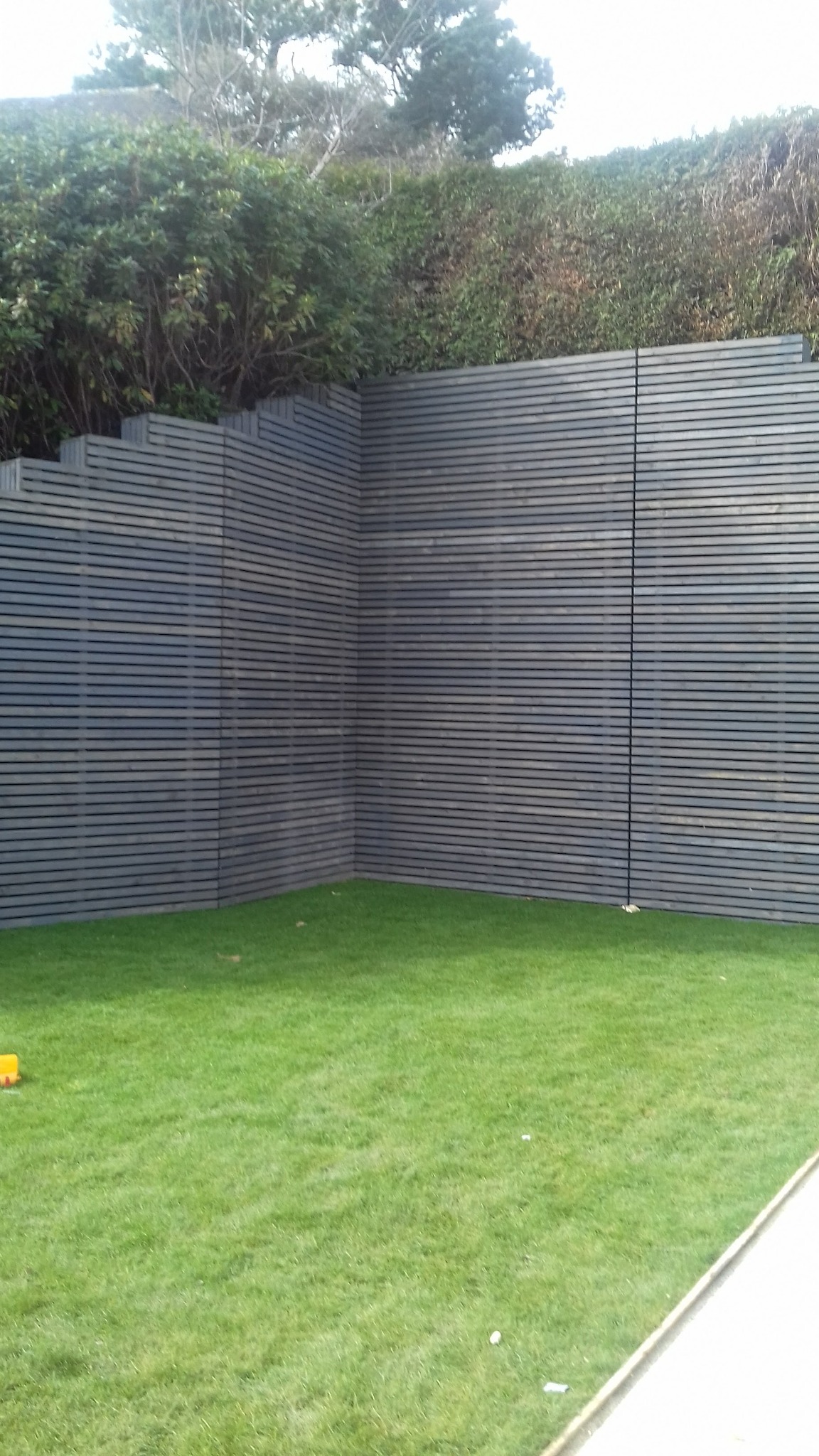 Nowoczesne ogrodzenie z poziomych, szarych desek, tworzące narożnik na tle zielonego trawnika i gęstych krzewów.