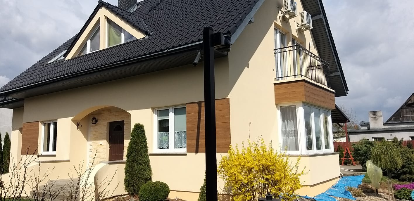 Jasna elewacja domu jednorodzinnego z balkonem, drewnianymi akcentami i czarną dachówką, ozdobiona kwitnącym krzewem forsycji.