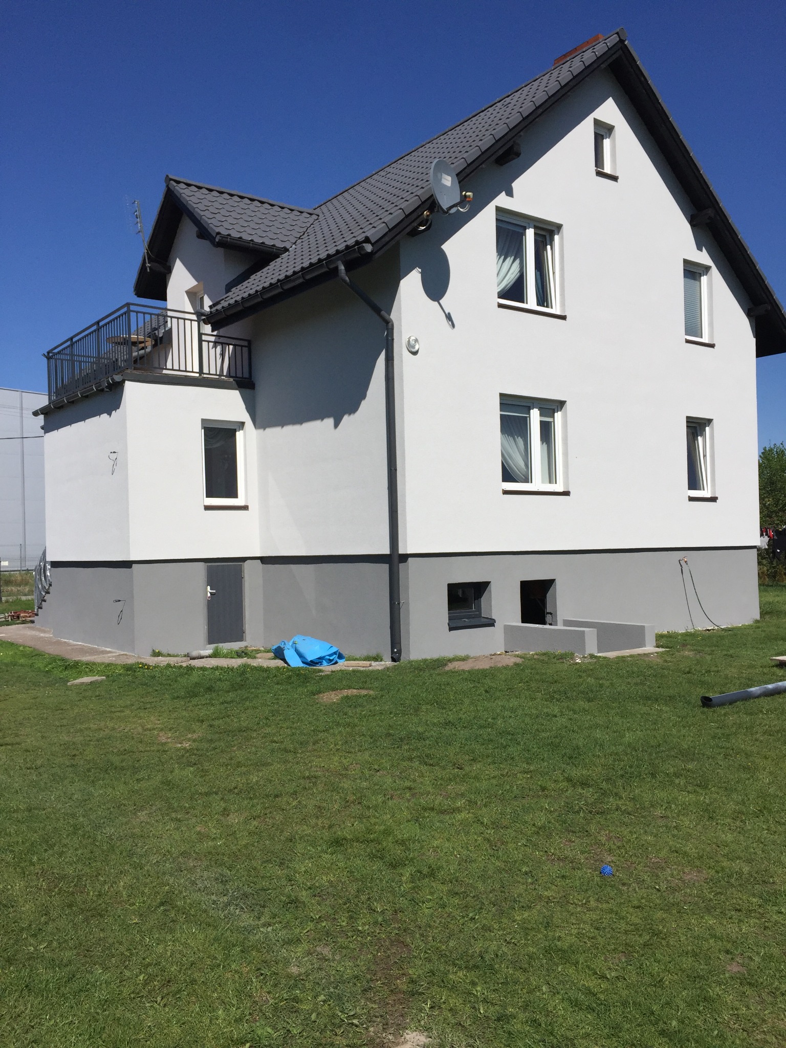 Dwupiętrowy dom z szarą elewacją, ciemnoszarą podmurówką, balkonem z czarną balustradą i szarym dachem, widziany z poziomu trawnika w słoneczny dzień.