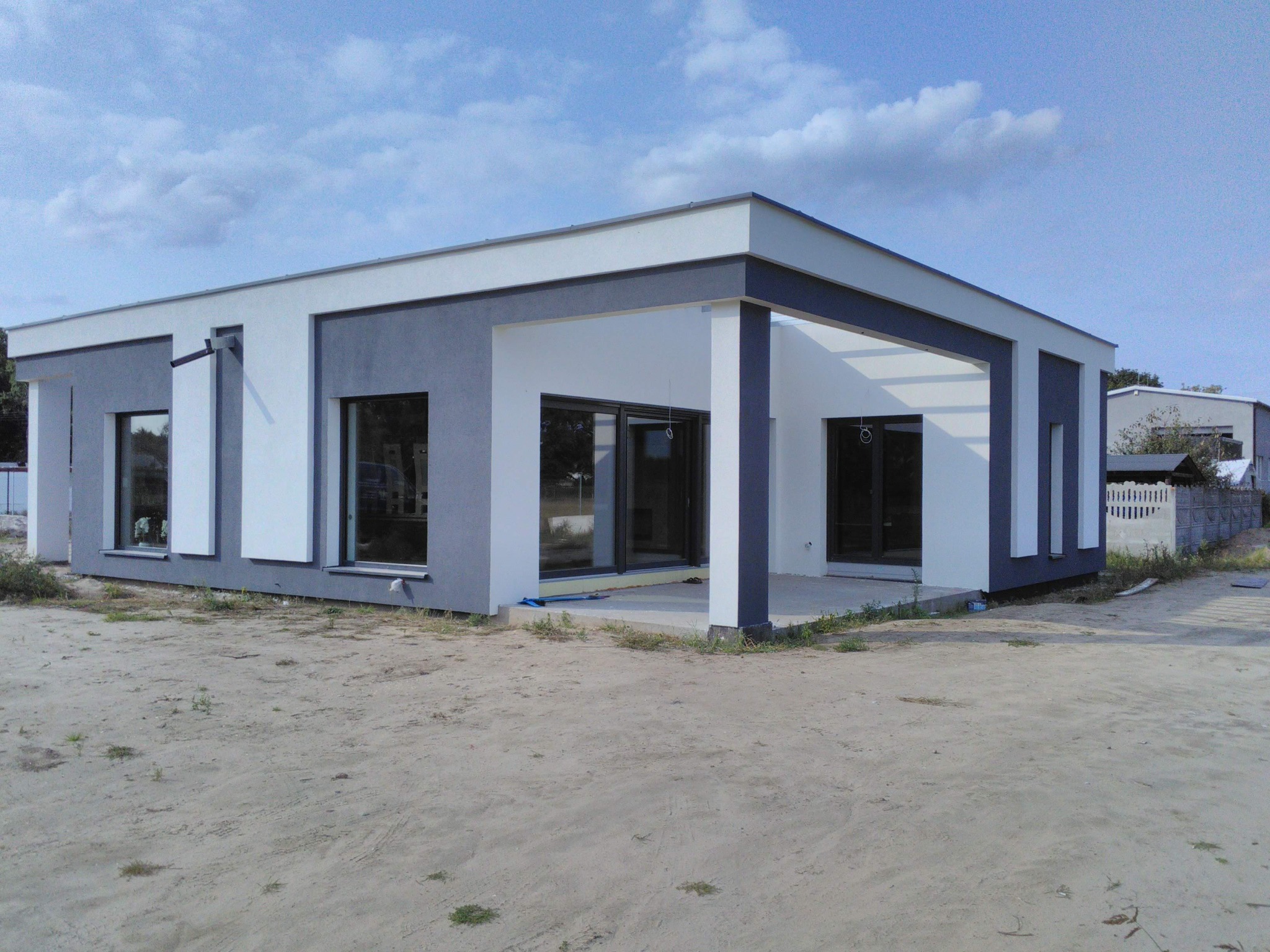 Nowoczesny dom jednorodzinny z płaskim dachem, wykończony w kolorach szarości i bieli, z dużymi przesuwnymi oknami tarasowymi i odkrytym zadaszeniem tarasu.