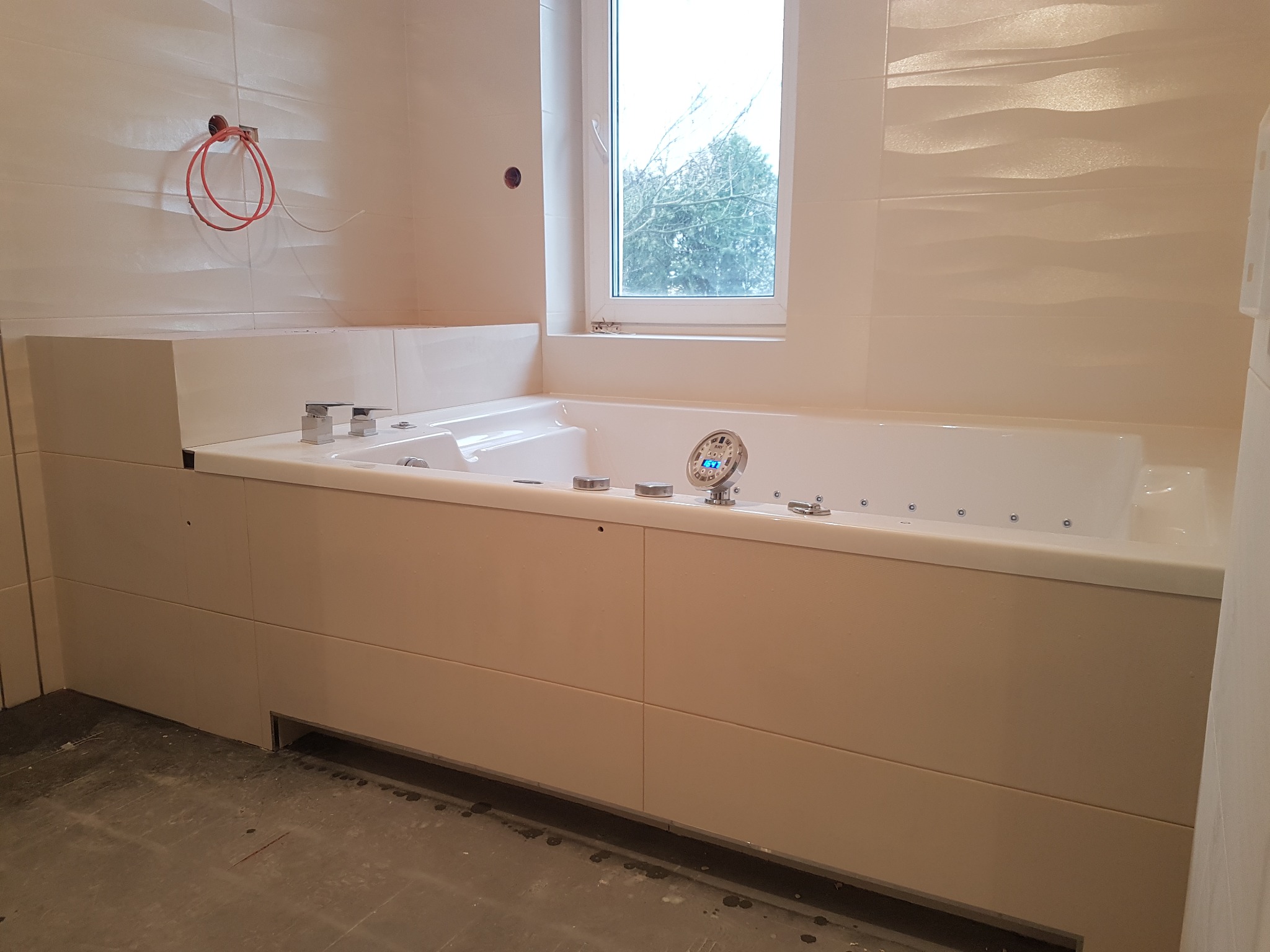 Nowoczesna wanna z hydromasażem w jasnej łazience z oknem. Widoczne niedokończone prace: wystające kable i surowa podłoga. Płytki ścienne z delikatnym wzorem.