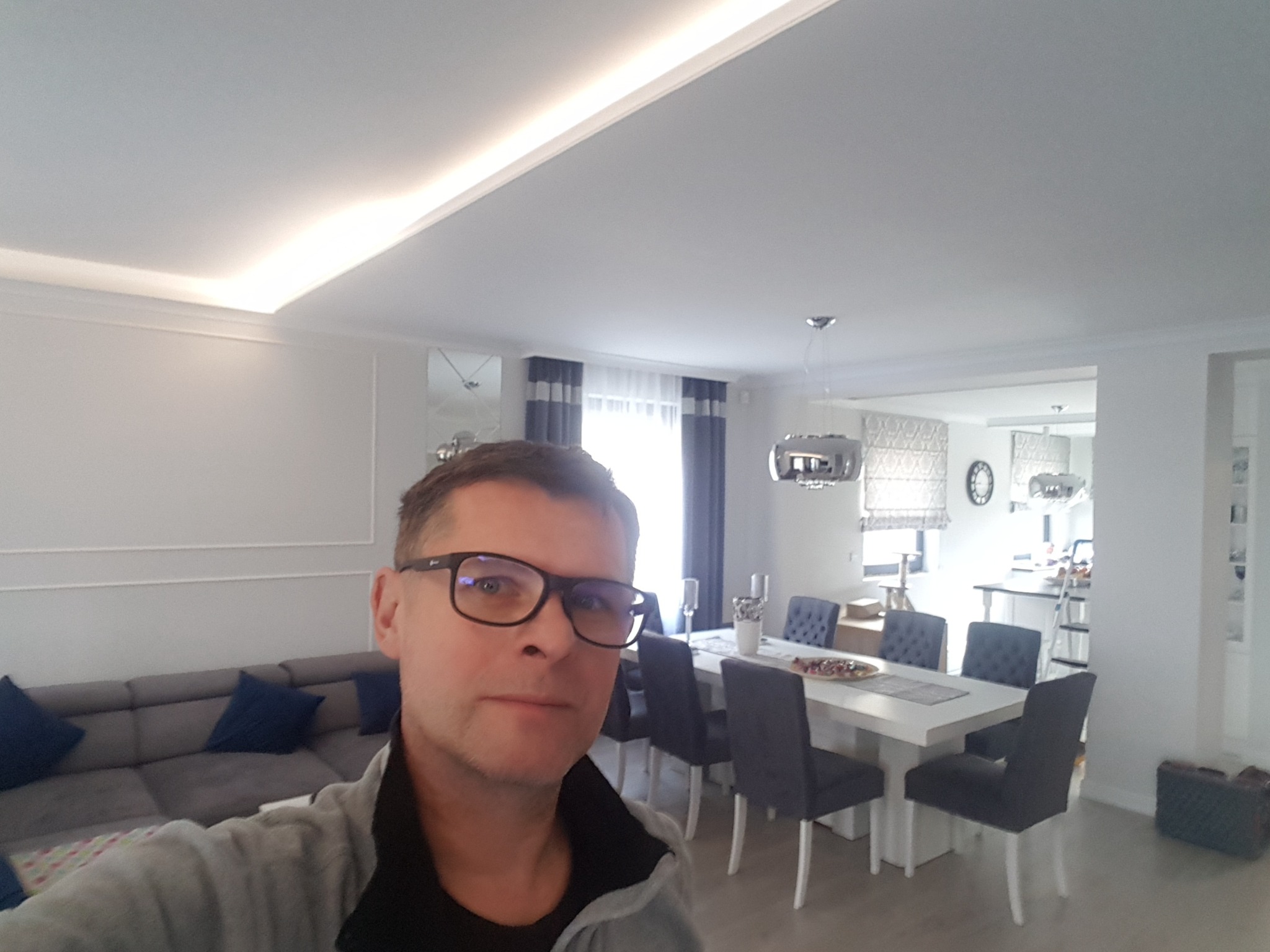 Jasne wnętrze po liftingu: salon z szarą sofą, biały stół jadalny z krzesłami i podświetlany sufit. Mężczyzna w okularach w kadrze.