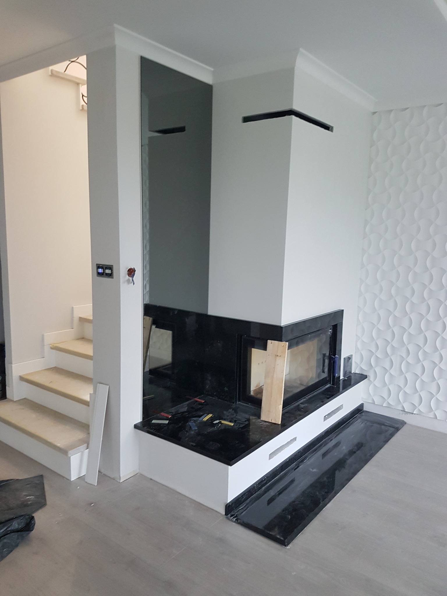 Wnętrze w trakcie wykańczania z nowoczesnym kominkiem z czarnym obramowaniem, białą obudową i dekoracyjną ścianą z wzorem 3D, widoczne schody z surowego drewna i narzędzia na obudowie kominka.