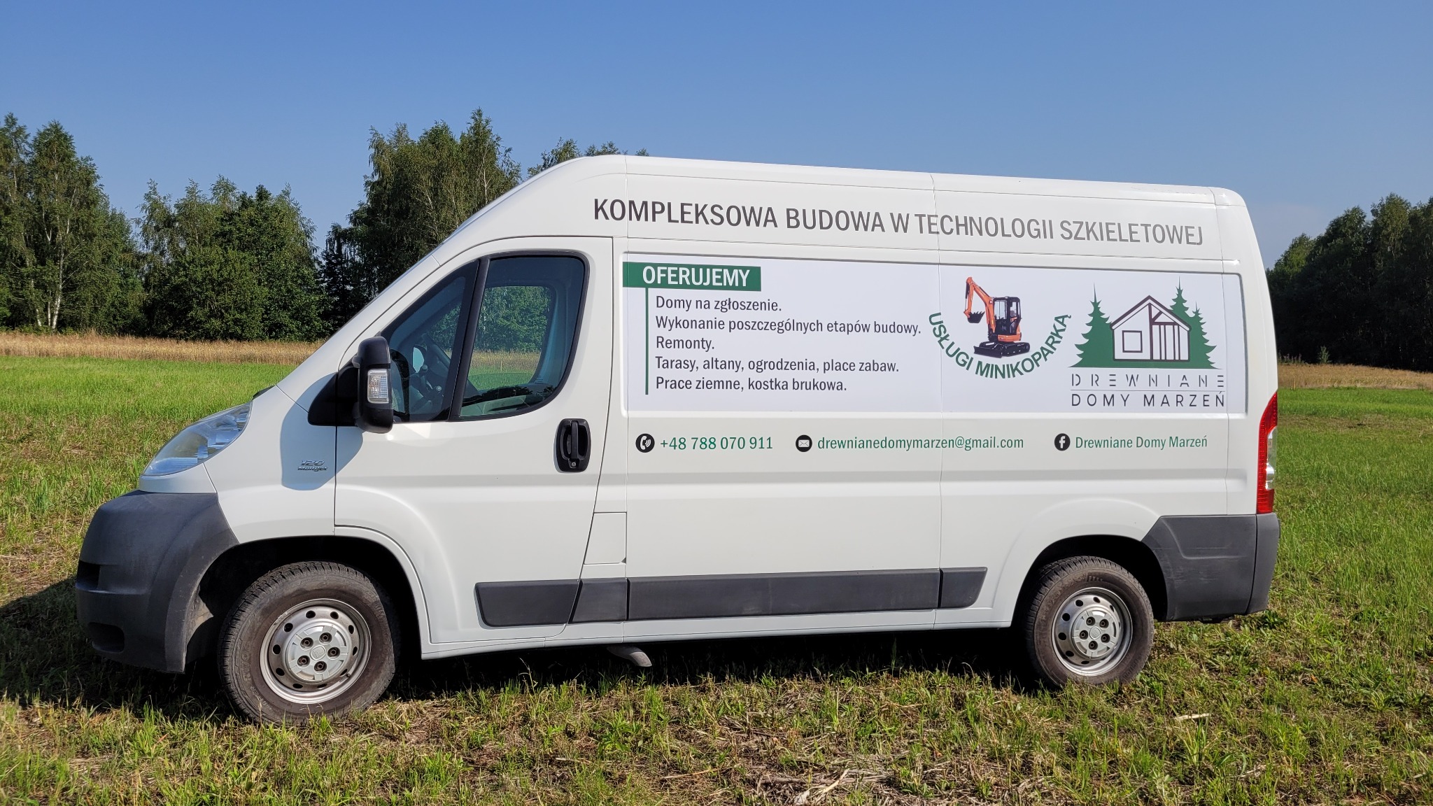 Biały samochód dostawczy z reklamą firmy budującej domy w technologii szkieletowej, zaparkowany na zielonej łące w słoneczny dzień.