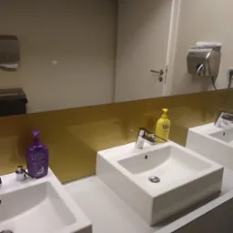 Umywalki w publicznej toalecie z dozownikami mydła Milla w kolorze fioletowym i żółtym, widoczne automatyczne suszarki do rąk i podajnik papieru.