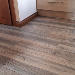 UNIFLOR - Nowa, jasnobrązowa podłoga z paneli imitujących drewno, ułożona wzdłużnie, widoczna przy listwach przypodłogowych i drzwiach z otworami wentylacyjnymi oraz szafkach kuchennych.