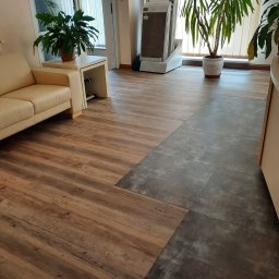 UNIFLOR - Połączenie dwóch rodzajów podłóg: paneli imitujących drewno i ciemnoszarych płytek, w jasnym wnętrzu z sofą i roślinami doniczkowymi.