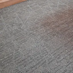 Szara wykładzina dywanowa z geometrycznym wzorem, widoczna listwa przypodłogowa i fragment mebla.