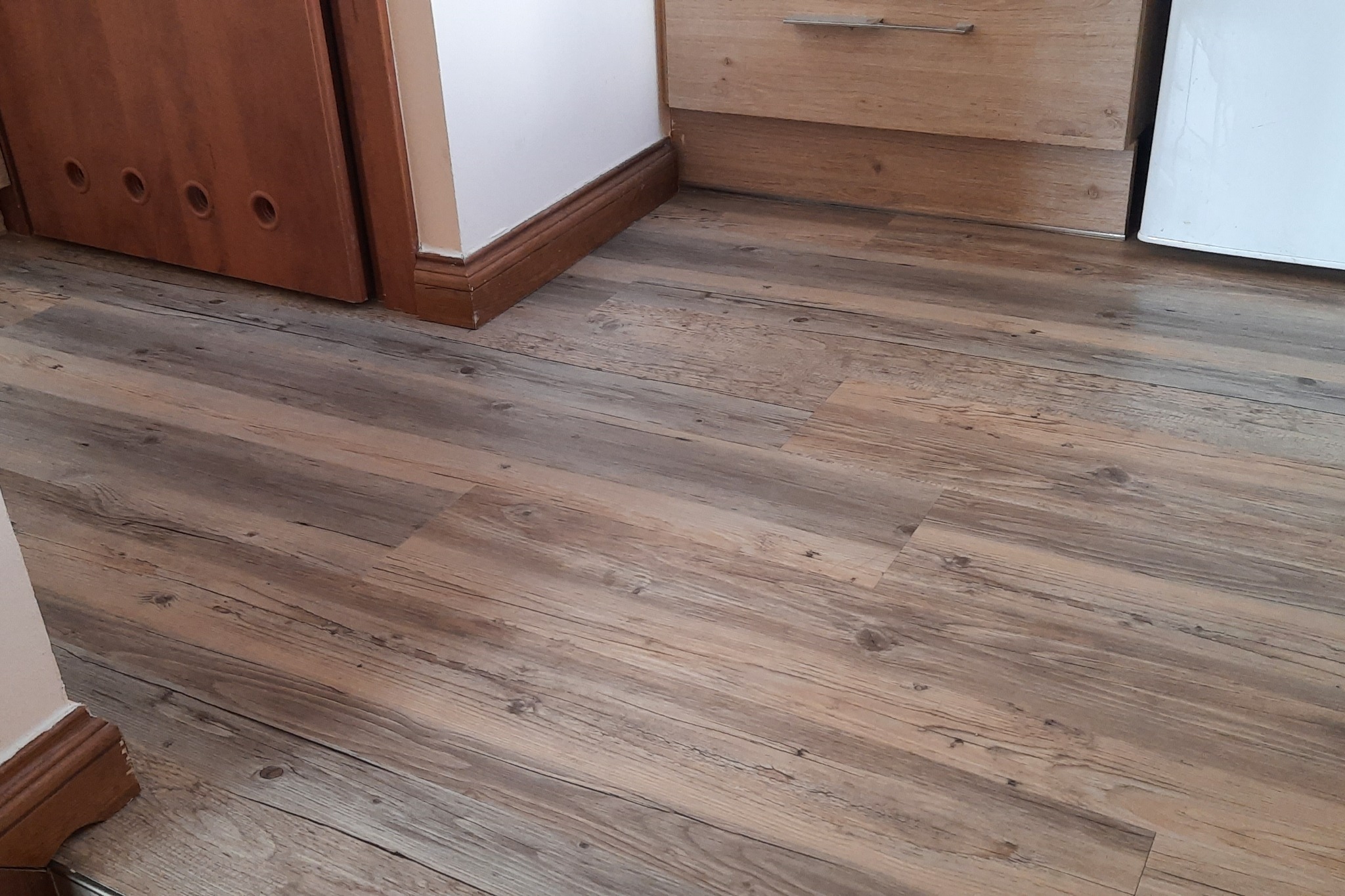 Nowa, jasnobrązowa podłoga z paneli imitujących drewno, ułożona wzdłużnie, widoczna przy listwach przypodłogowych i drzwiach z otworami wentylacyjnymi oraz szafkach kuchennych.