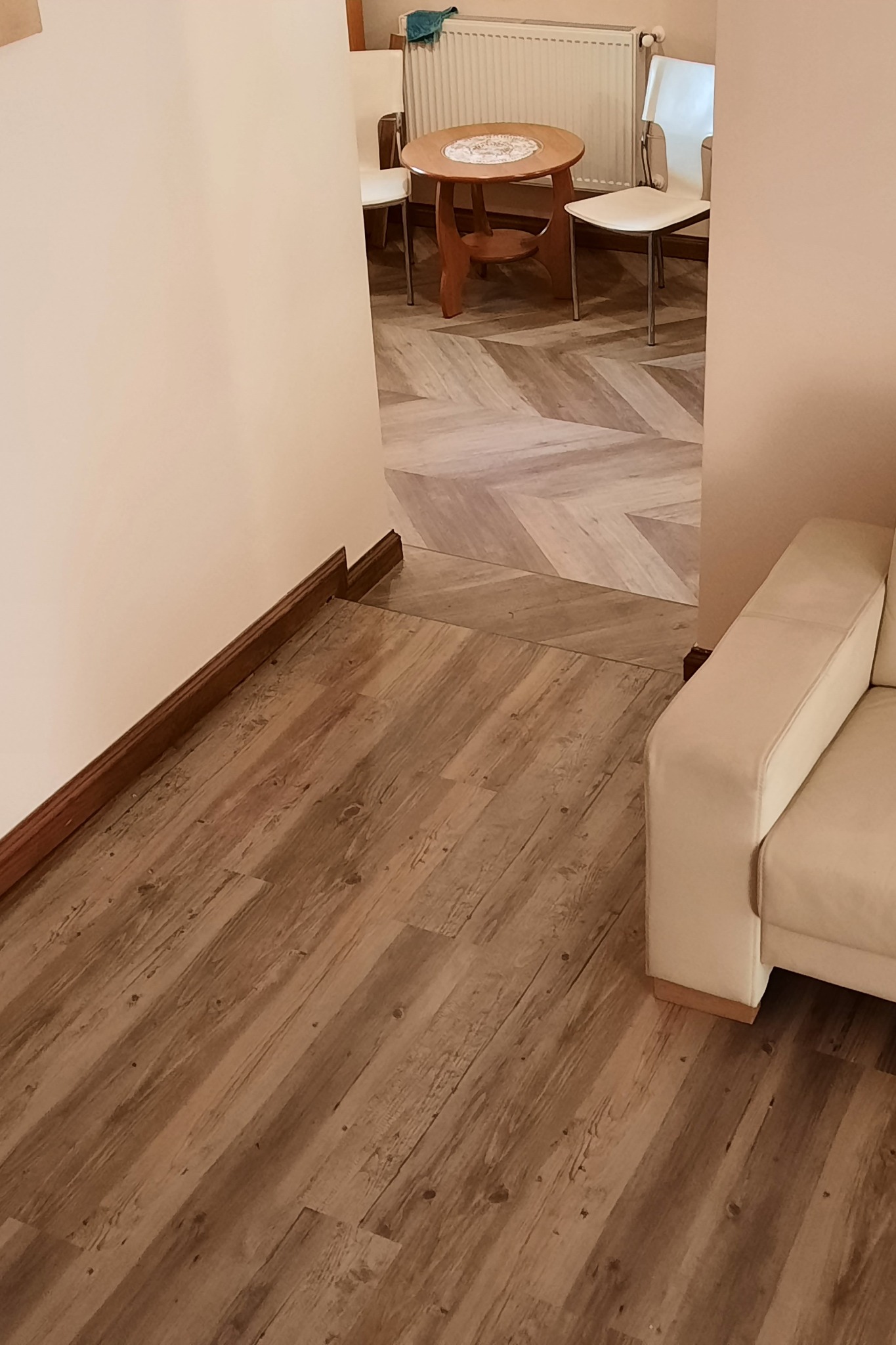 Ułożona pod kątem drewnopodobna podłoga przechodzi w jodełkę w dalszej części pokoju, widoczny fragment kremowej sofy i listwa przypodłogowa.