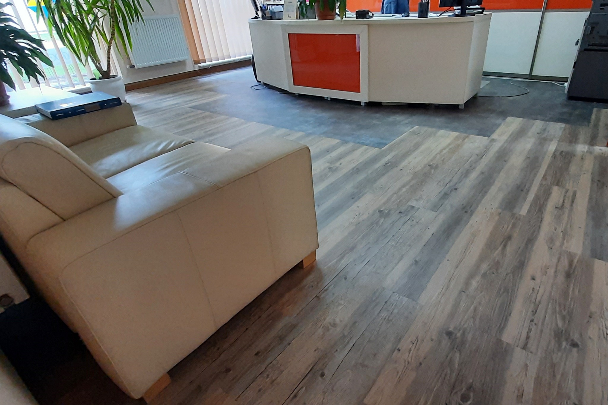 Wnętrze biura z jasną sofą i wykładziną imitującą drewno, widoczna recepcja w tle.