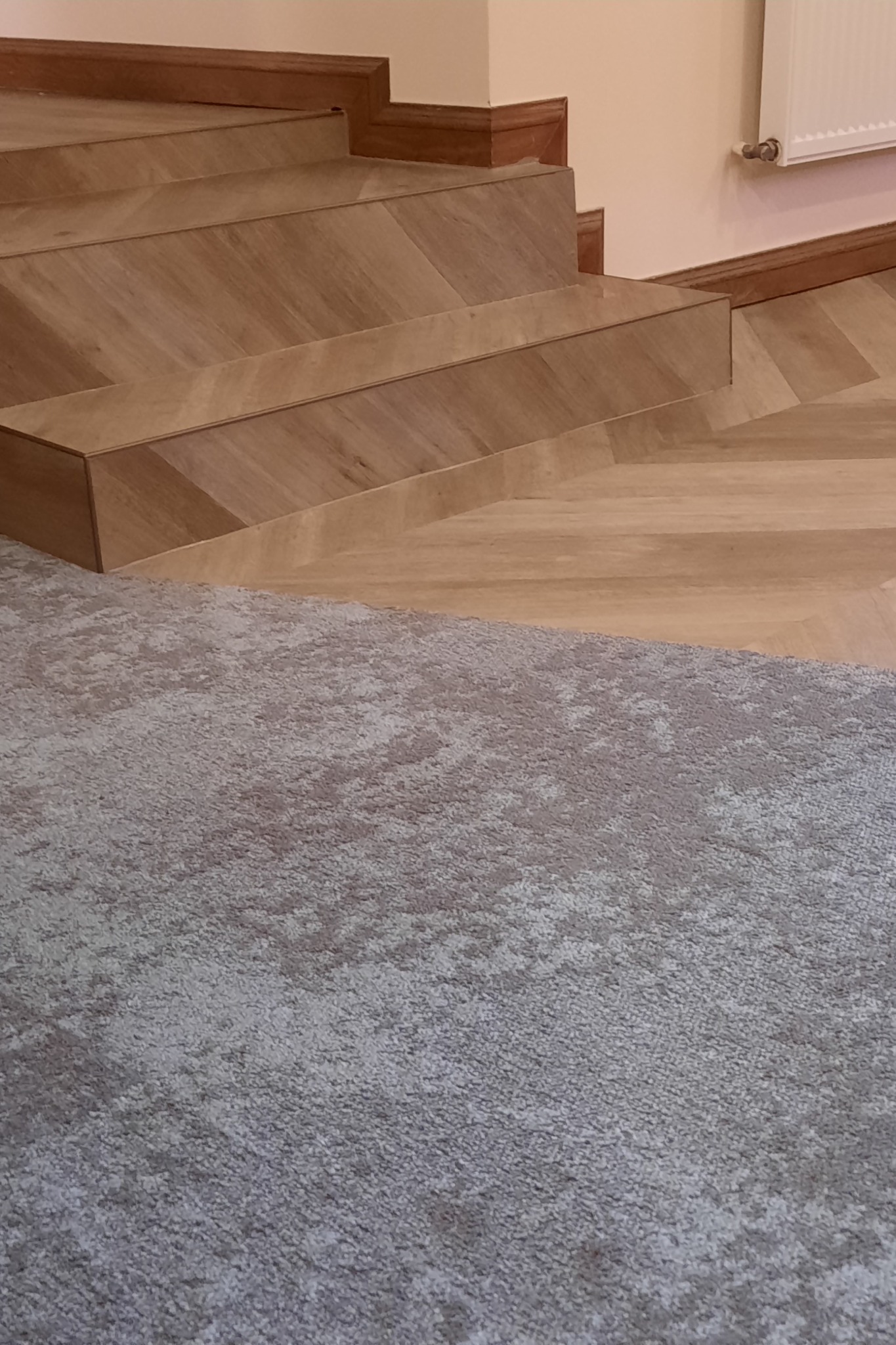 Fragment schodów wykończonych panelami ułożonymi w jodełkę, sąsiadujący z szarą wykładziną dywanową o delikatnym wzorze.