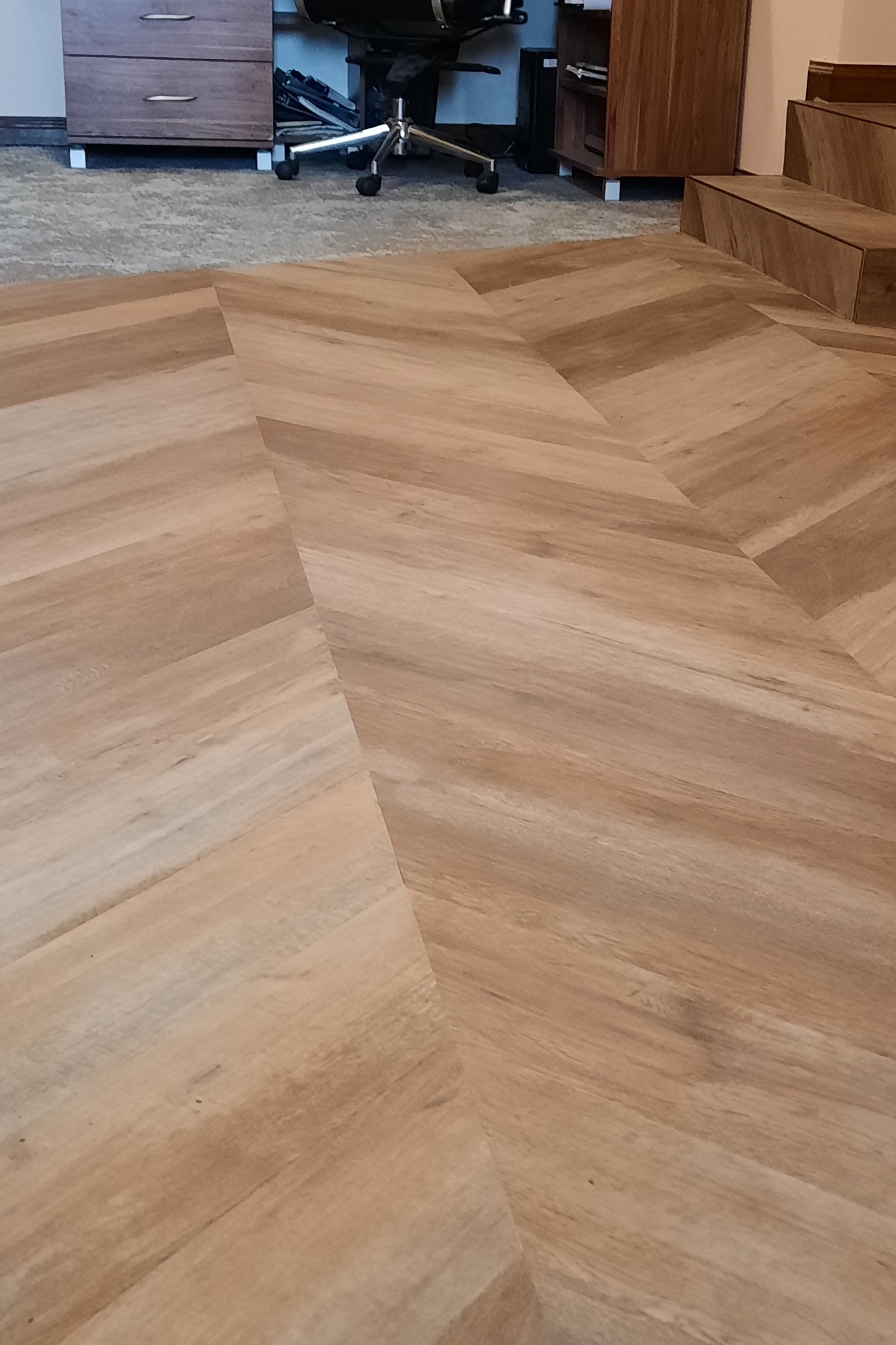 Jasna, drewnopodobna podłoga ułożona we wzór jodełki francuskiej, widoczne fragmenty mebli i biurowego krzesła w tle.