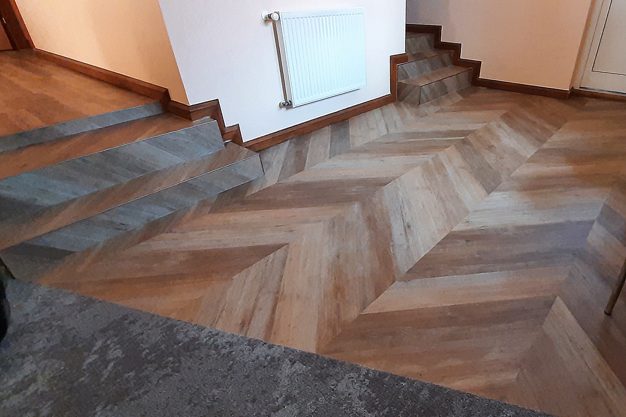 Ułożona w jodełkę jasna podłoga z paneli laminowanych, z widocznymi schodami i grzejnikiem na ścianie.