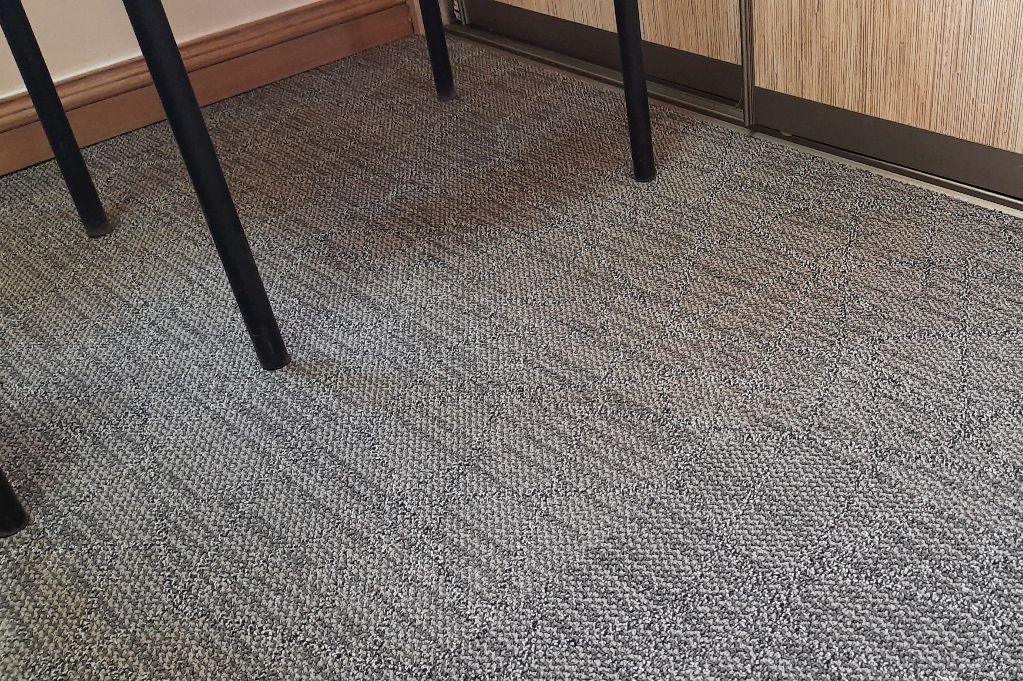 Szara, wzorzysta podłoga pokryta wykładziną dywanową, widoczne nogi czarnego krzesła i fragment drewnianej listwy przypodłogowej oraz szafy przesuwnej.