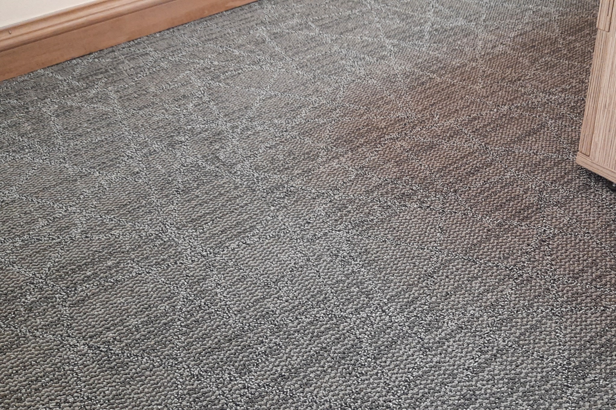 Szara wykładzina dywanowa z geometrycznym wzorem, widoczna listwa przypodłogowa i fragment mebla.