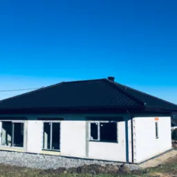 Nowo wybudowany dom jednorodzinny z czarną dachówką, widok z zewnątrz na tle błękitnego nieba, stan surowy otwarty, okna i drzwi zamontowane.