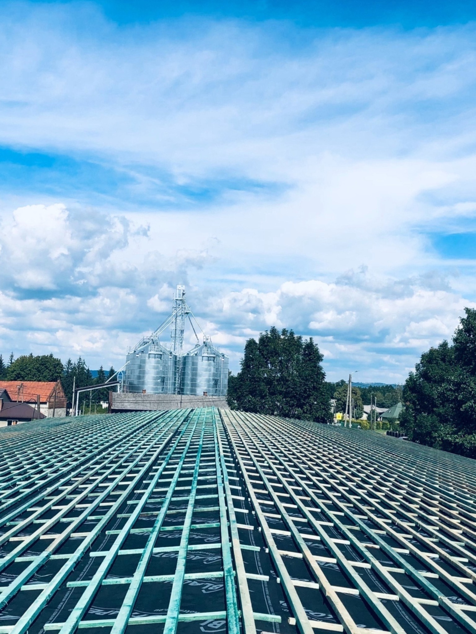 Wykonywanie więźby dachowej na czarnej membranie dachowej, w tle zabudowania gospodarcze i błękitne niebo z chmurami.