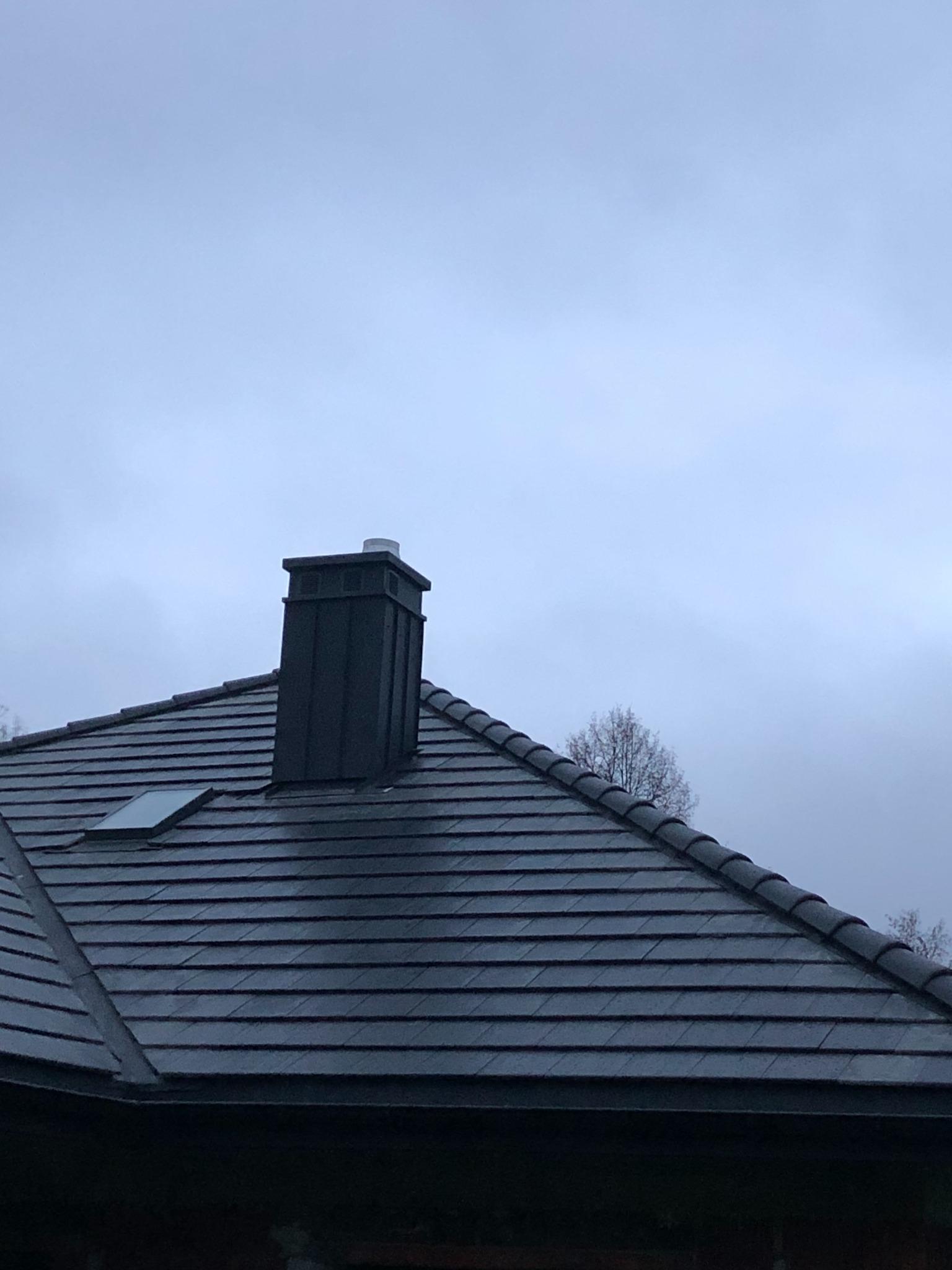 Ciemna dachówka ceramiczna z oknem dachowym i kominem na tle pochmurnego nieba.