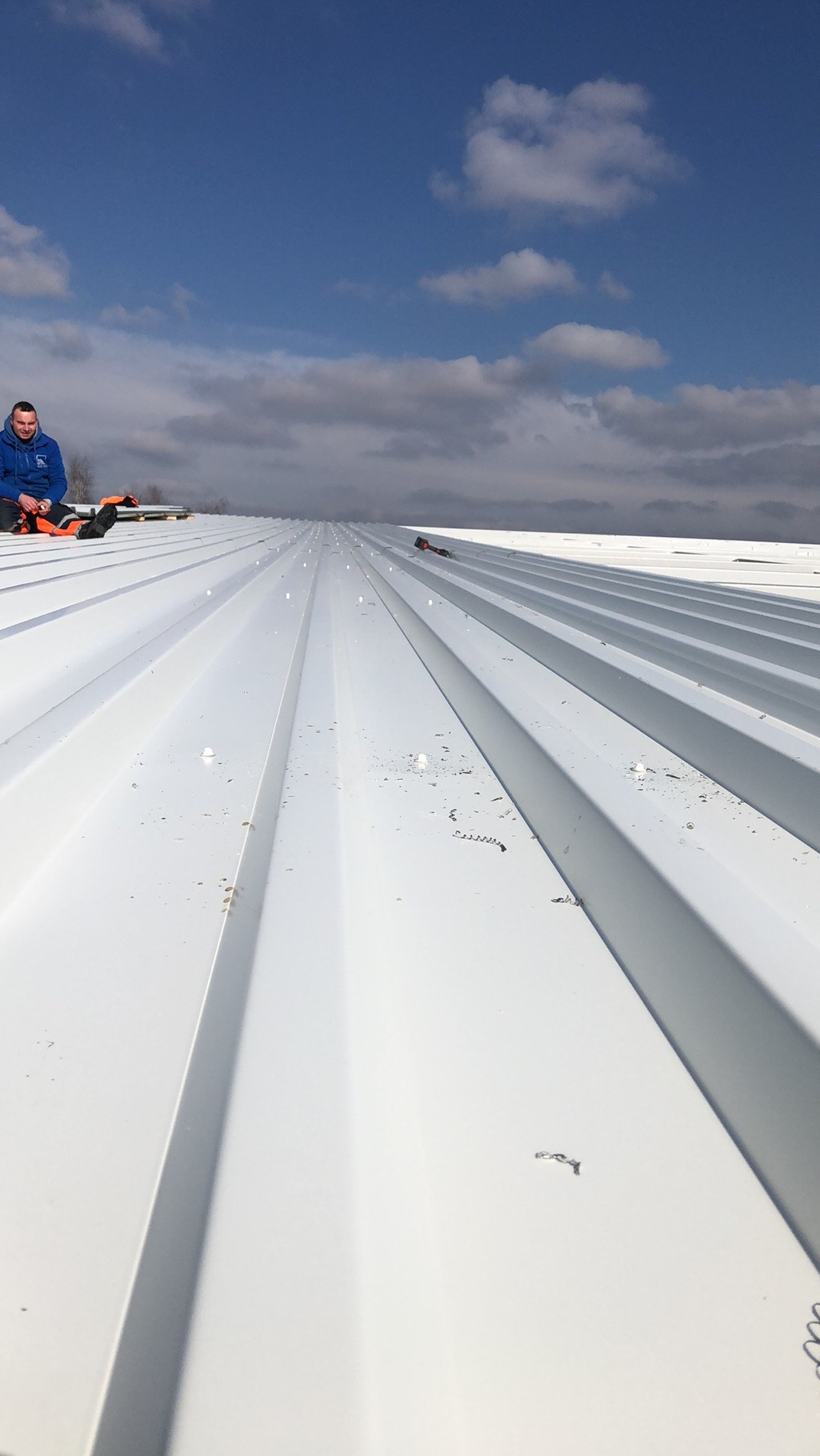 Nowy, biały dach z blachy trapezowej, widoczne wkręty i wióry po wierceniu, dekarz siedzi na dachu w niebieskiej bluzie i pomarańczowych spodniach, błękitne niebo z chmurami w tle.