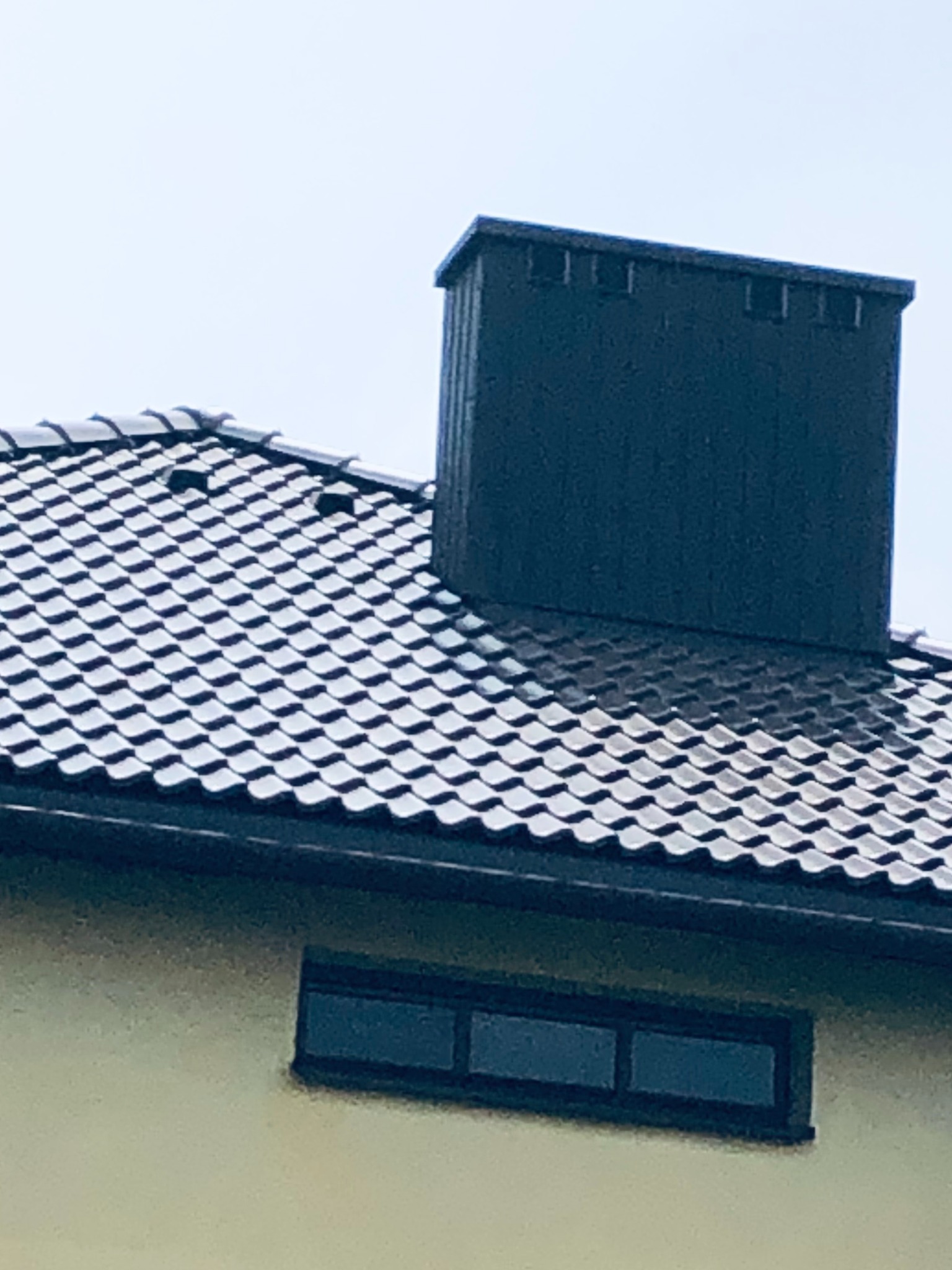 Szczegółowy widok dachu z ciemnej dachówki ceramicznej, z kominem w kolorze grafitowym i fragmentem ściany budynku z małym, prostokątnym oknem.