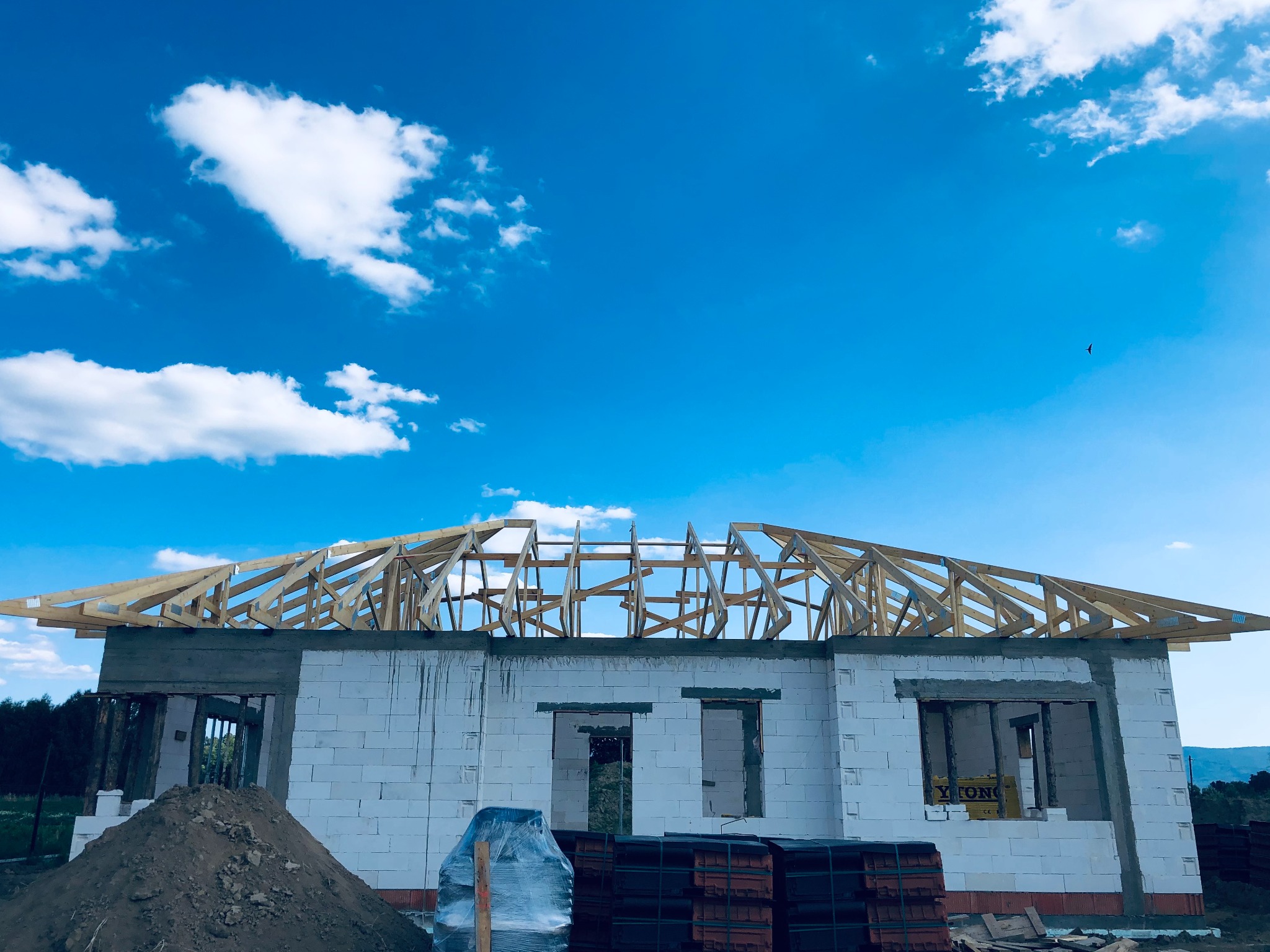Budowa domu jednorodzinnego: surowy stan z widoczną więźbą dachową na tle błękitnego nieba z chmurami, palety z dachówką obok ściany z betonu komórkowego.