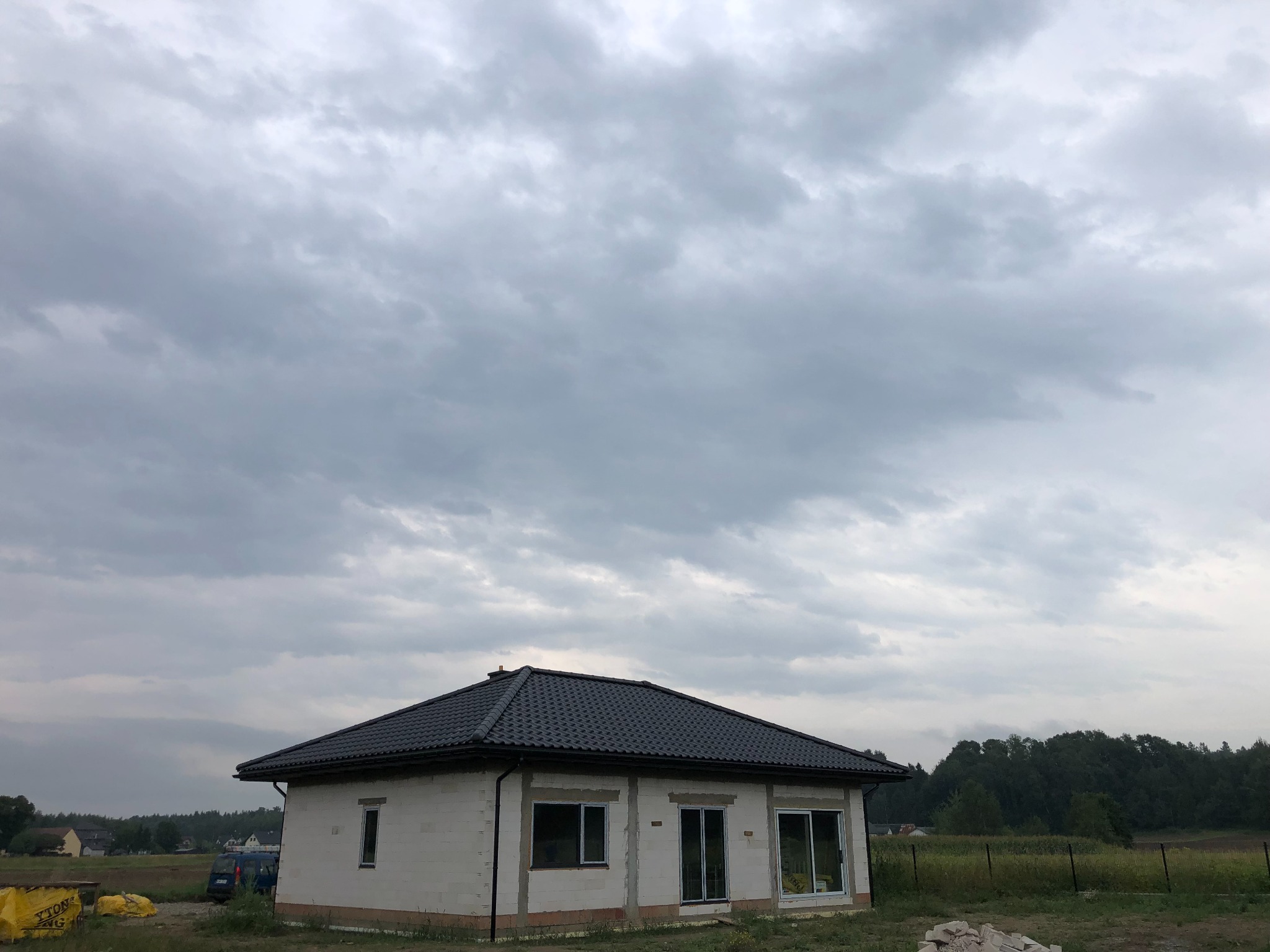 Niedokończony dom jednorodzinny z szarym dachem pod pochmurnym niebem, widoczne worki z materiałami budowlanymi na trawie.