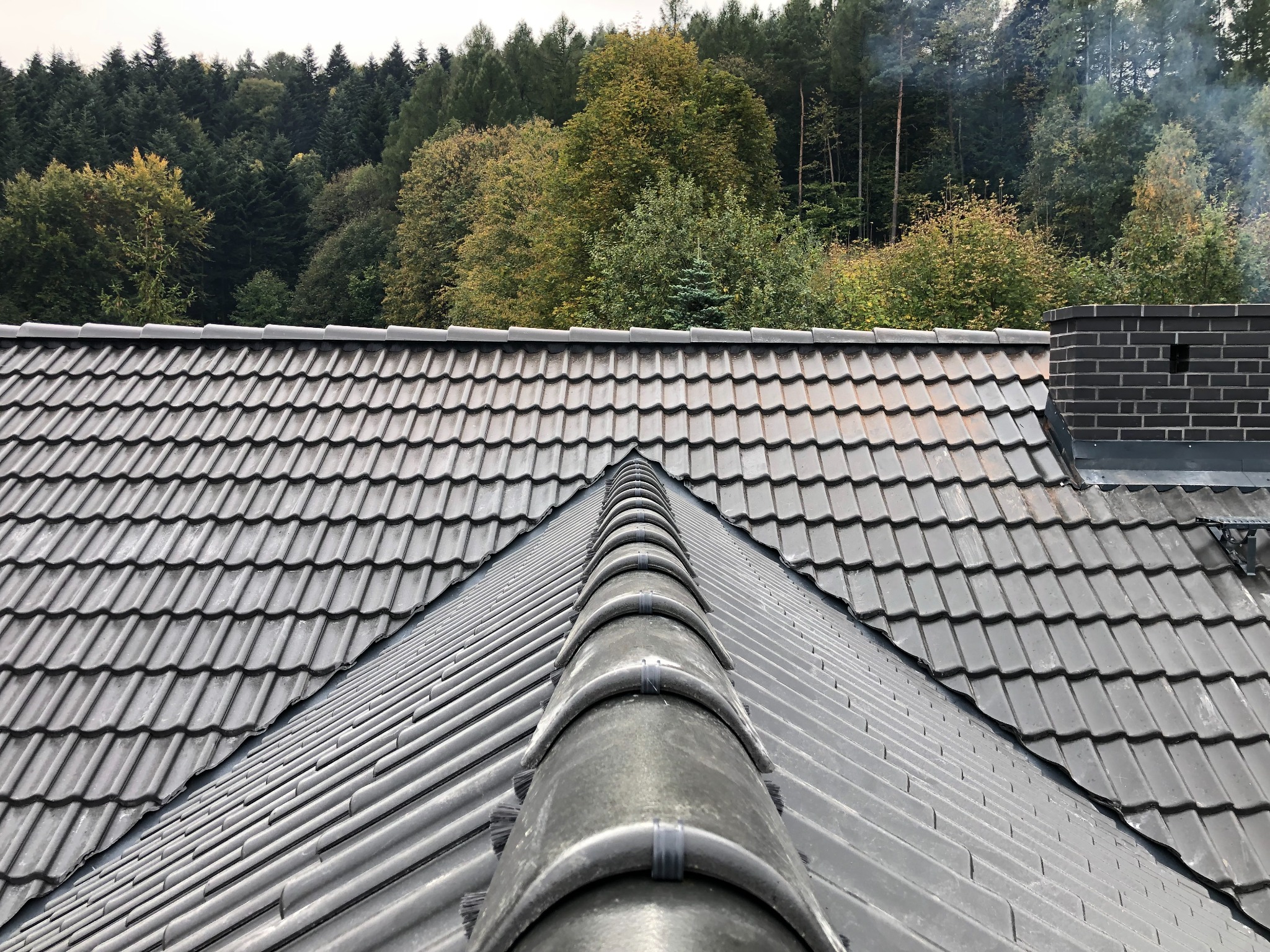 Nowoczesny dach pokryty szarą dachówką ceramiczną, widok z góry na kalenicę, w tle las i komin z cegły.