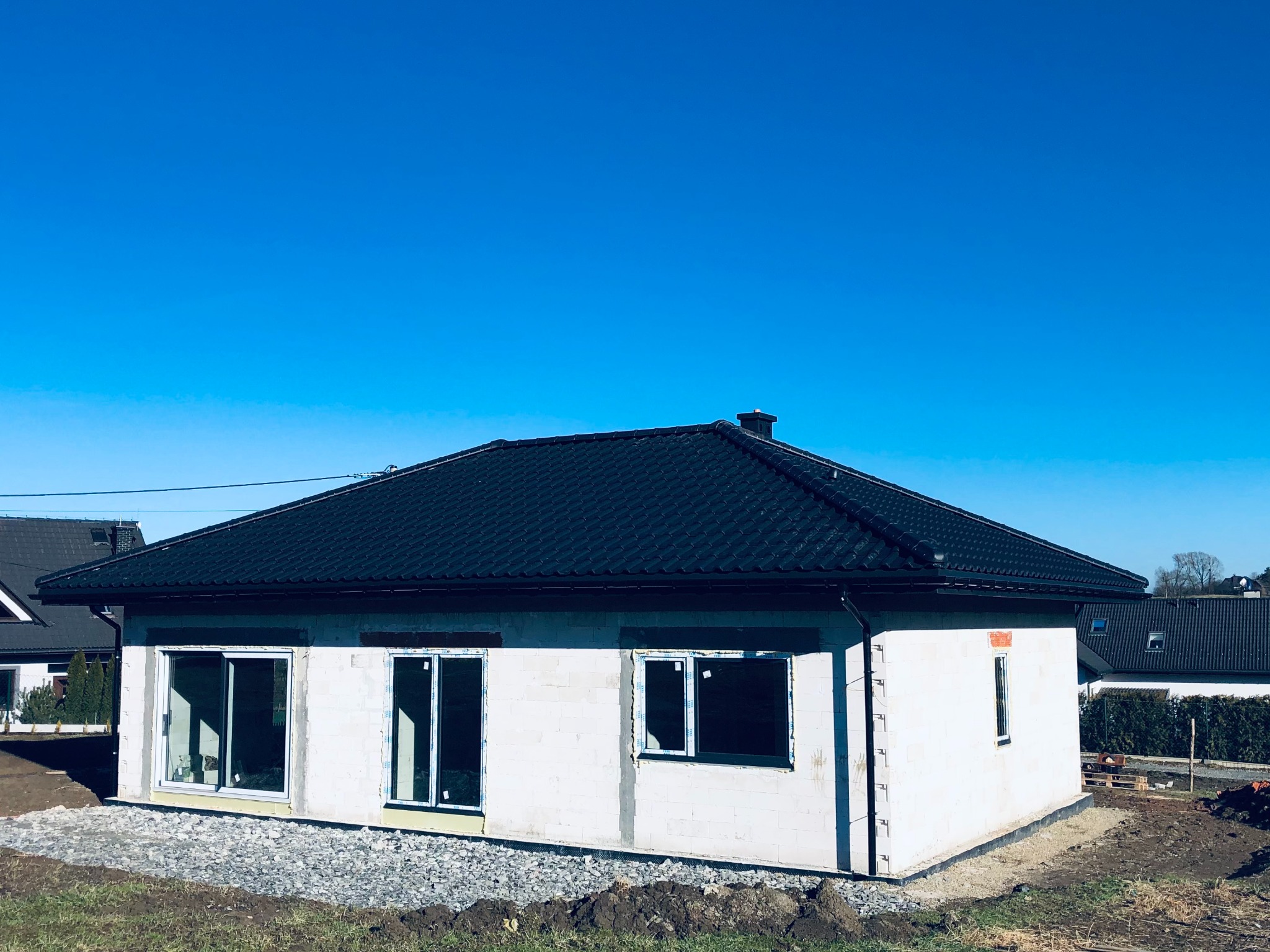 Nowo wybudowany dom jednorodzinny z czarną dachówką, widok z zewnątrz na tle błękitnego nieba, stan surowy otwarty, okna i drzwi zamontowane.