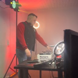 DJ Dandus - Daniel Piekarczyk Usługi Muzyczne - Mężczyzna w bluzie ustawia sprzęt DJ-ski Numark Mixtrack Pro FX na stole, oświetlenie sceniczne w tle, w tym lampy LED i ring light.
