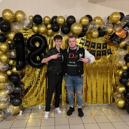 DJ Dandus - Daniel Piekarczyk Usługi Muzyczne - Dwóch młodych mężczyzn pozuje na tle złotej ścianki z balonów w kolorach czarnym i złotym, dekoracji z okazji 18 urodzin i napisu 'Happy Birthday'.