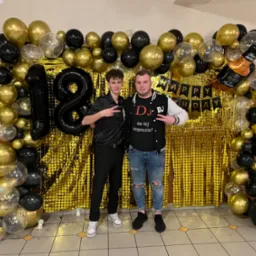 Dwóch młodych mężczyzn pozuje na tle złotej ścianki z balonów w kolorach czarnym i złotym, dekoracji z okazji 18 urodzin i napisu 'Happy Birthday'.