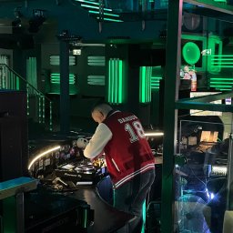 DJ Dandus - Daniel Piekarczyk Usługi Muzyczne - DJ w czerwonej kurtce miksuje muzykę na konsoli Pioneer DJ w klubie z zielonym oświetleniem LED, widoczny napis 'ENERGY 2000'