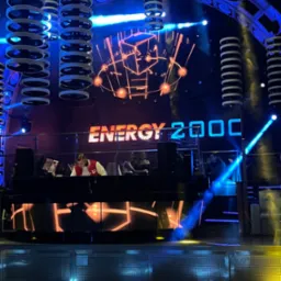 DJ w czerwono-białej kurtce za konsoletą na scenie klubu Energy 2000, oświetlonej niebieskimi i pomarańczowymi światłami, z dekoracjami sufitowymi w kształcie pierścieni.