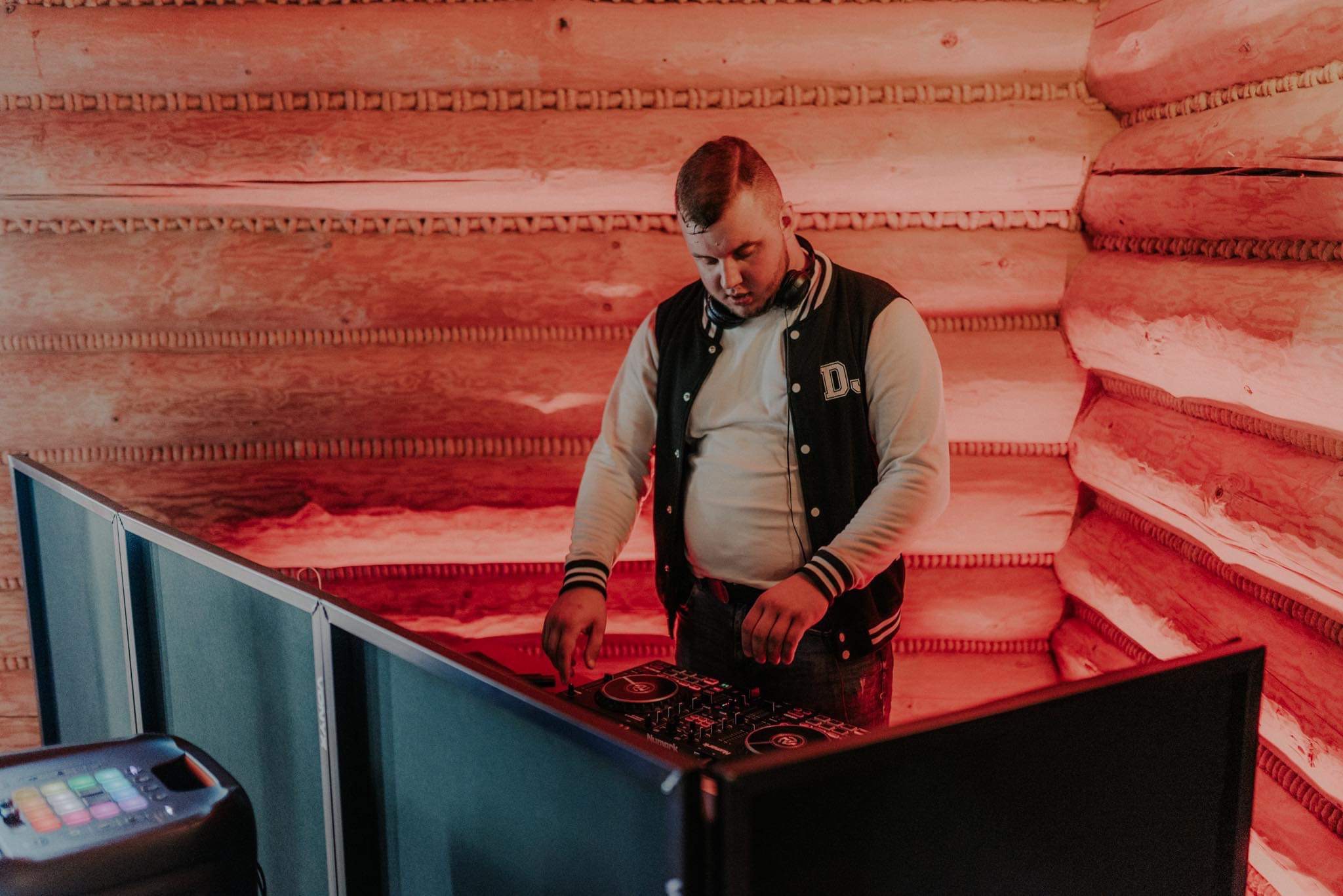 DJ w bomberce z literą D miksuje muzykę na konsoli Numark przed drewnianą ścianą z bali, oświetloną czerwonym światłem.