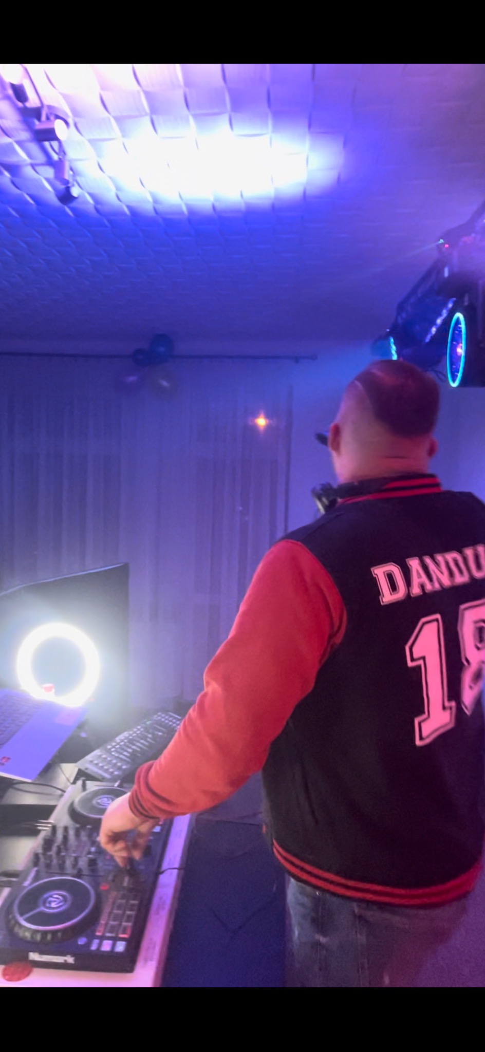 DJ w kurtce 'Dandu 18' obsługuje mikser Numark w niebieskim oświetleniu, widoczny laptop z ring light w tle.