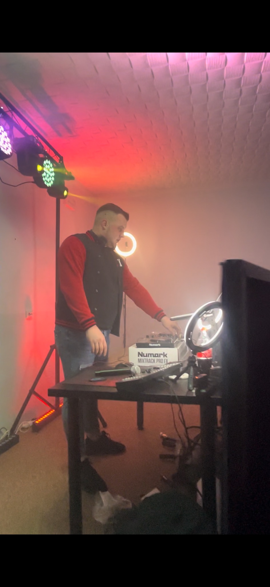 Mężczyzna w bluzie ustawia sprzęt DJ-ski Numark Mixtrack Pro FX na stole, oświetlenie sceniczne w tle, w tym lampy LED i ring light.
