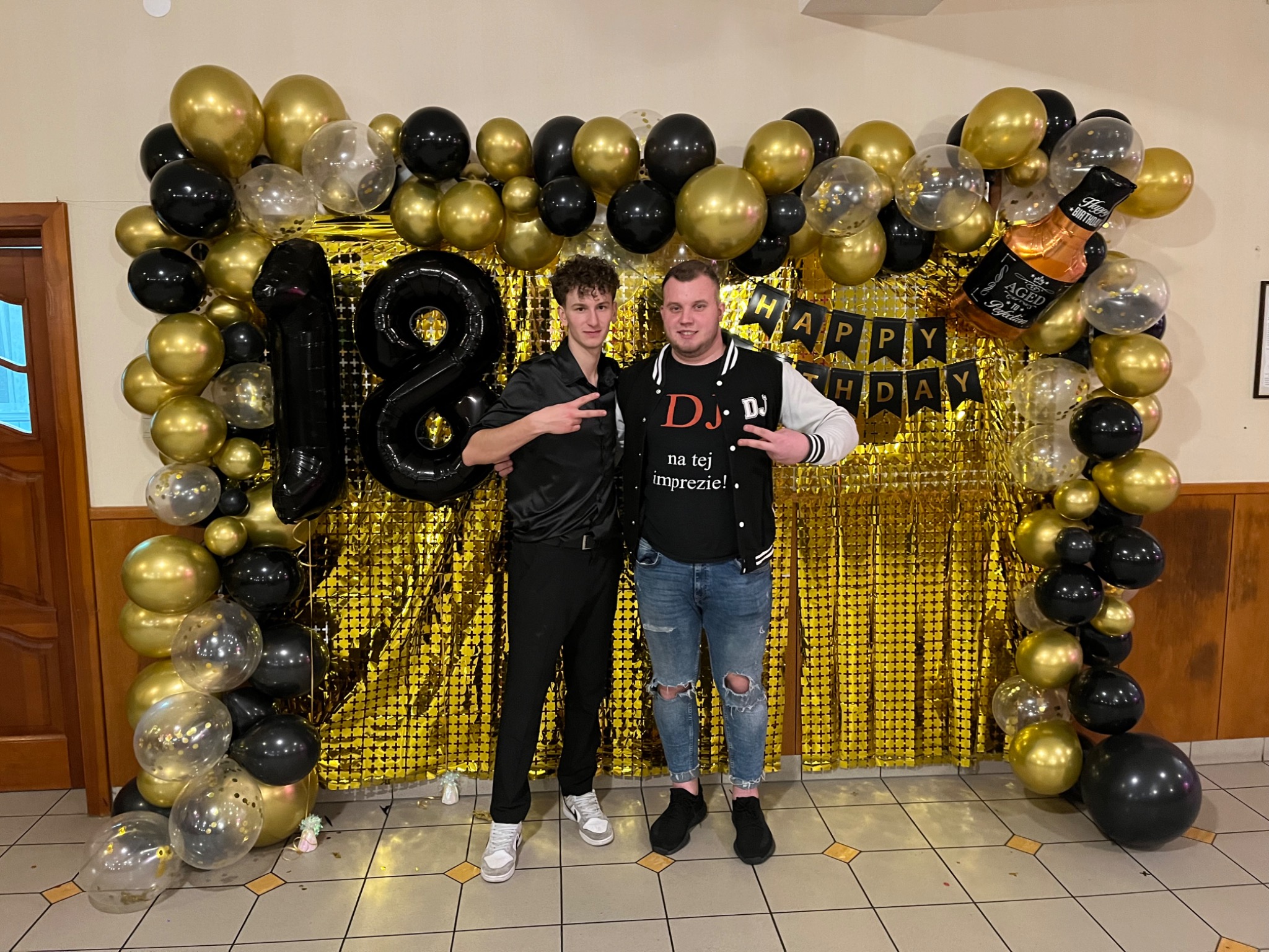 Dwóch młodych mężczyzn pozuje na tle złotej ścianki z balonów w kolorach czarnym i złotym, dekoracji z okazji 18 urodzin i napisu 'Happy Birthday'.