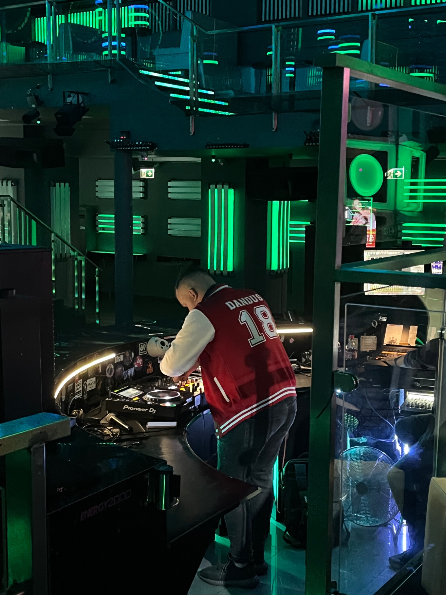 DJ w czerwonej kurtce miksuje muzykę na konsoli Pioneer DJ w klubie z zielonym oświetleniem LED, widoczny napis 'ENERGY 2000'