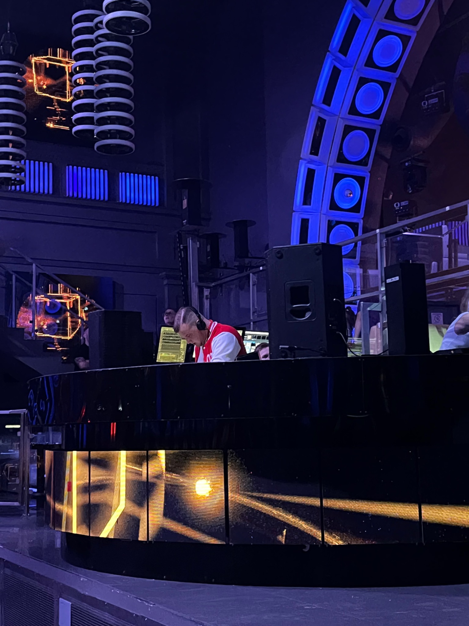 DJ w czerwonej kurtce za podświetlonym panelem, na scenie z dekoracyjnym oświetleniem i nagłośnieniem.