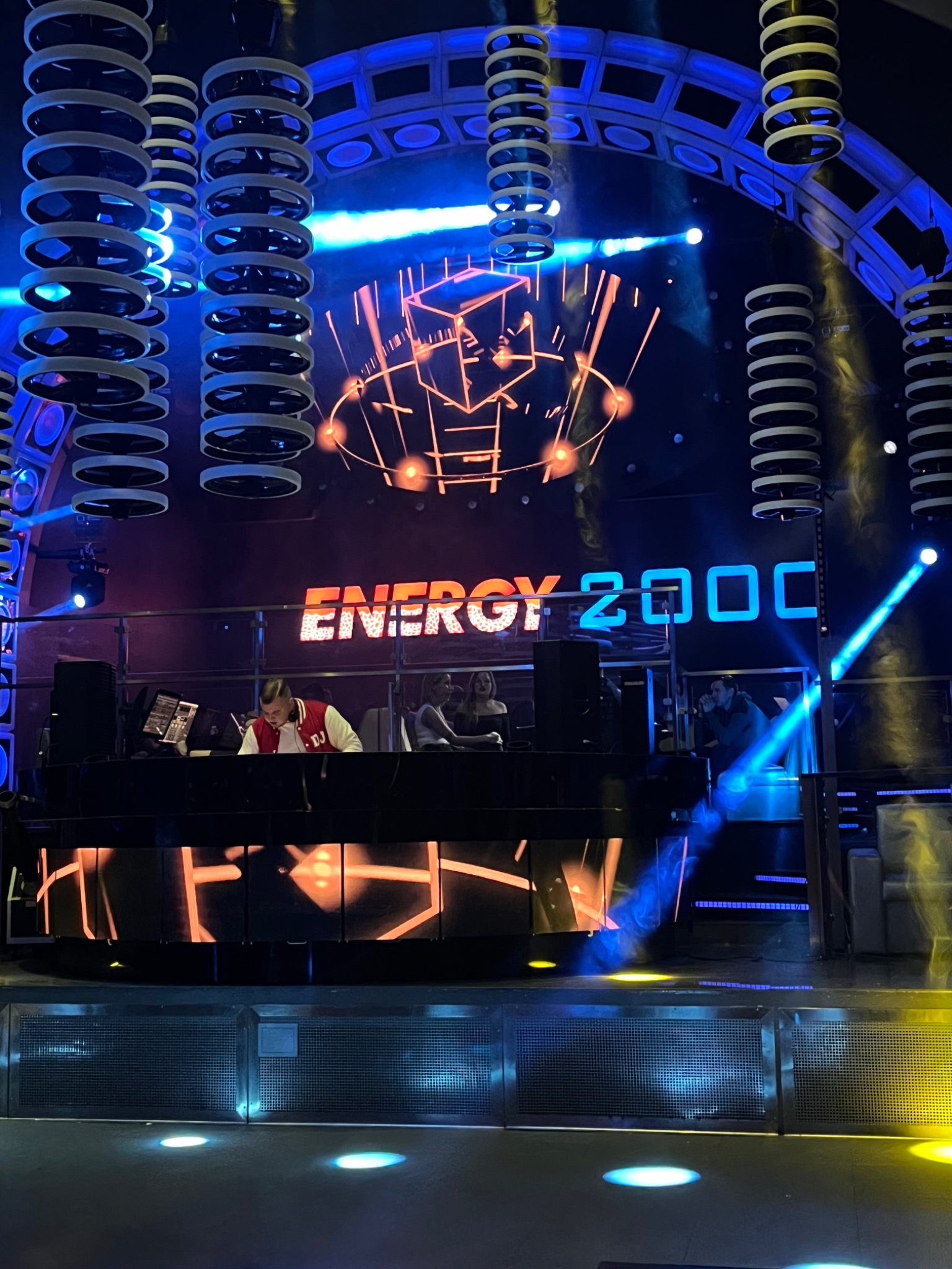DJ w czerwono-białej kurtce za konsoletą na scenie klubu Energy 2000, oświetlonej niebieskimi i pomarańczowymi światłami, z dekoracjami sufitowymi w kształcie pierścieni.