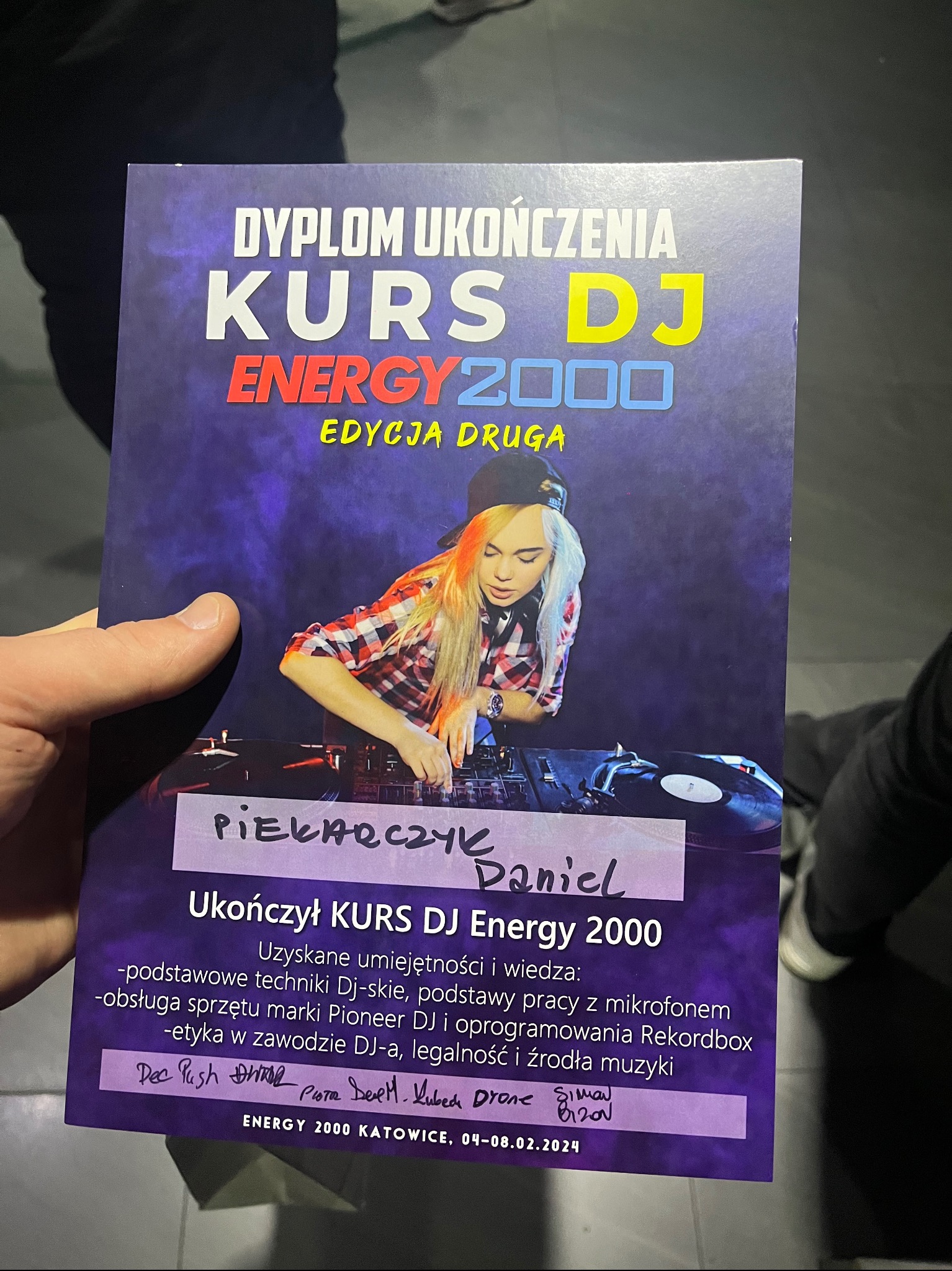 Dyplom ukończenia kursu DJ Energy 2000, edycja druga, trzymany w dłoni, z wizerunkiem DJ-ki przy sprzęcie.