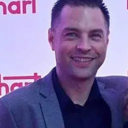 Portret uśmiechniętego mężczyzny w szarej marynarce na tle czerwonej ściany z białymi napisami.