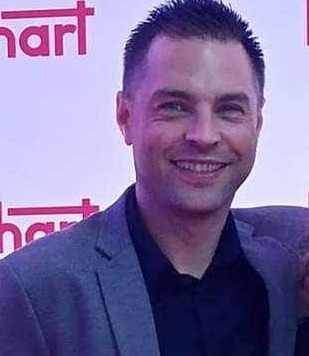 Portret uśmiechniętego mężczyzny w szarej marynarce na tle czerwonej ściany z białymi napisami.