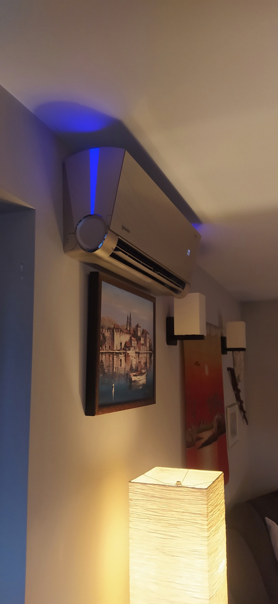 Klimatyzator ścienny z niebieską linią LED, zamontowany w pomieszczeniu mieszkalnym, pod nim obraz z motywem miejskim i lampa podłogowa.
