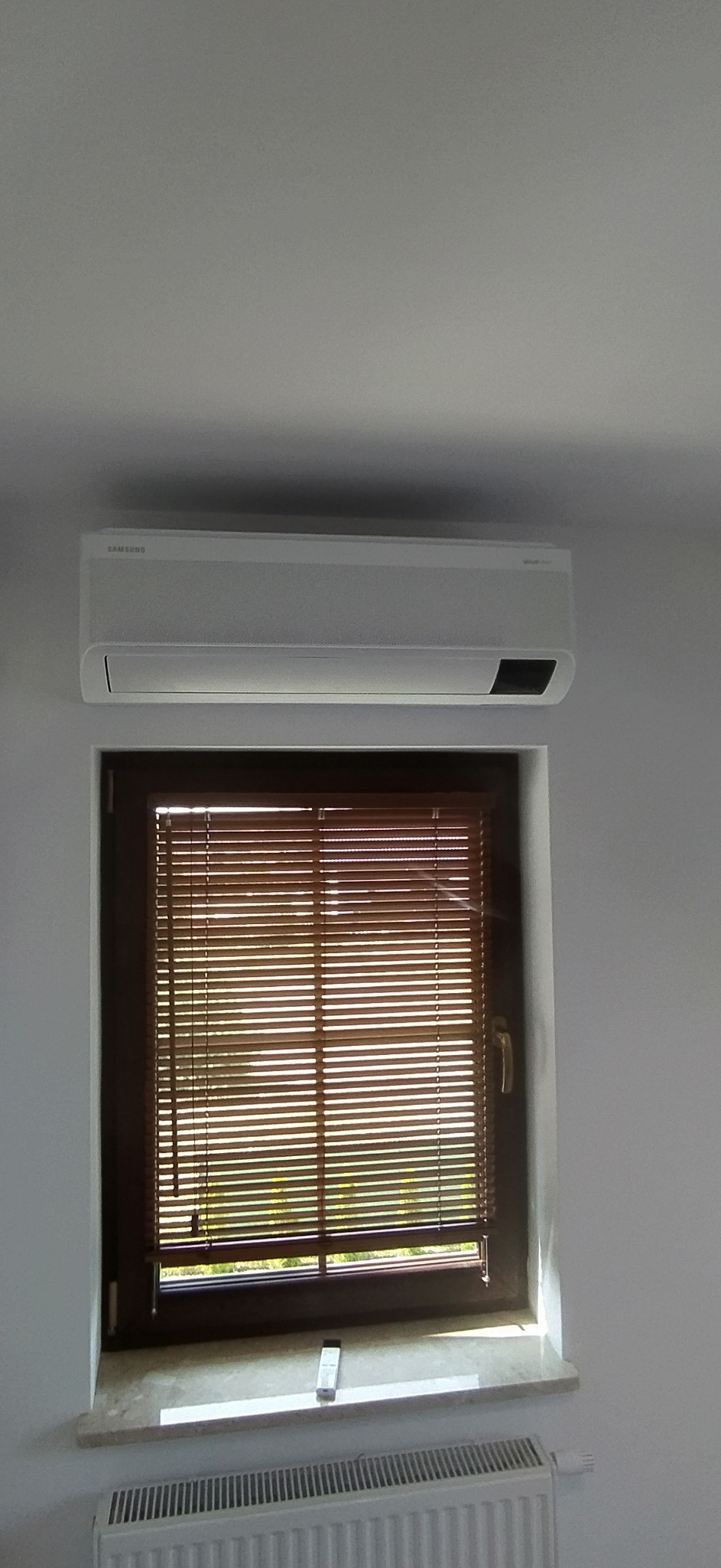Biały klimatyzator Samsung zamontowany nad oknem z drewnianymi żaluzjami w jasnym pomieszczeniu, widoczny biały grzejnik pod oknem i pilot do klimatyzacji na parapecie.