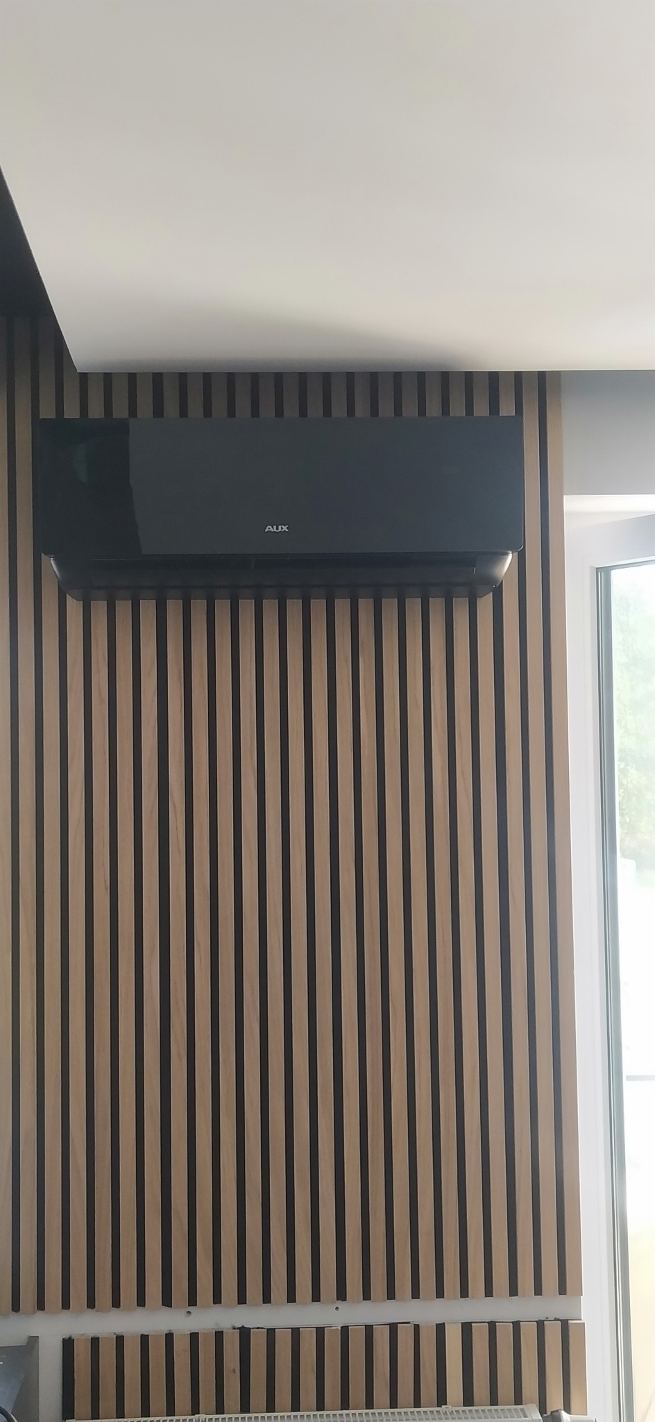 Czarny klimatyzator marki AUX zamontowany na ścianie z dekoracyjnych paneli drewnianych, widoczne okno w tle.