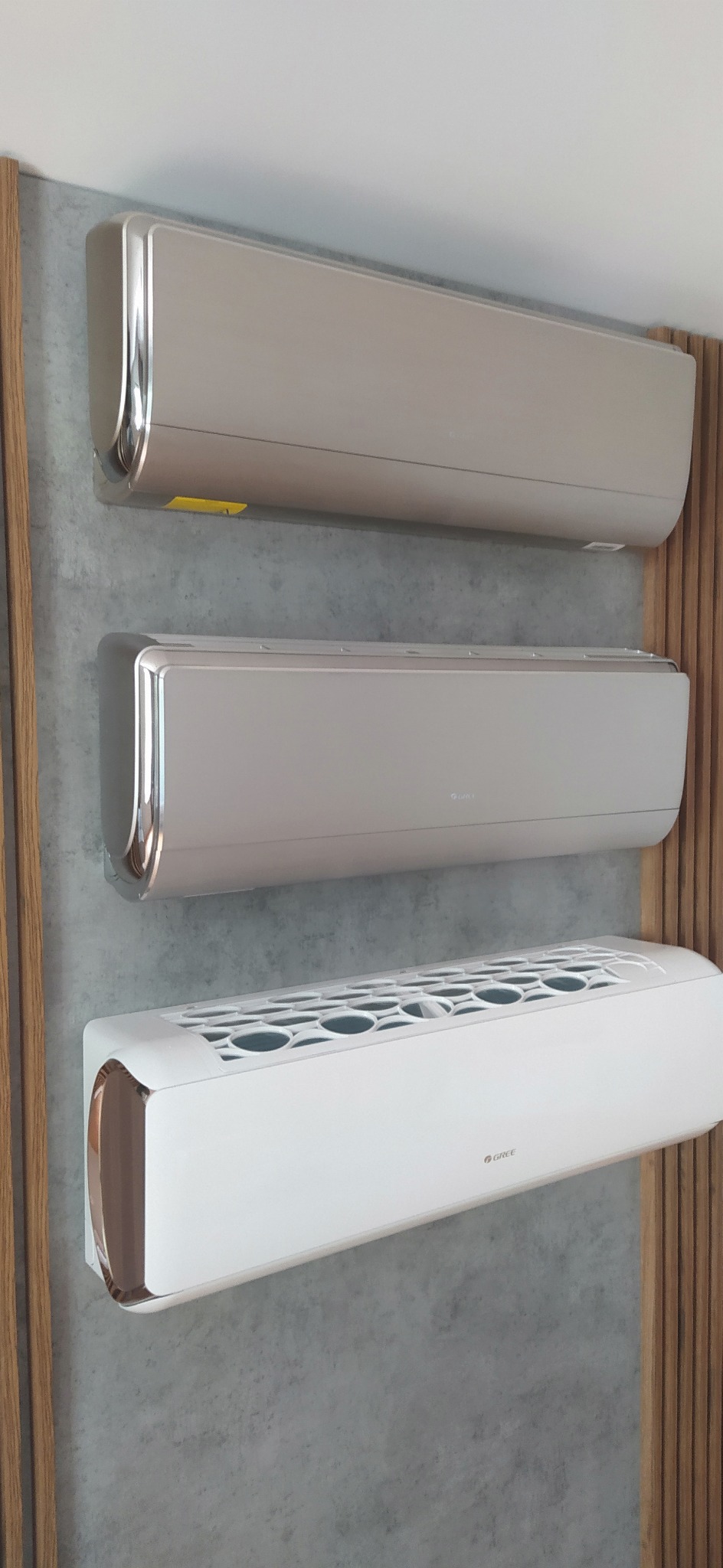 Trzy klimatyzatory ścienne różnych kolorów (biały, szary, beżowy) zamontowane obok siebie na szarej ścianie z drewnianą dekoracją.